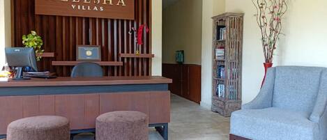 A reception area with a wooden desk, two cushioned stools, a grey armchair, a bookshelf, and a sign that reads "ALEESHA VILLAS".