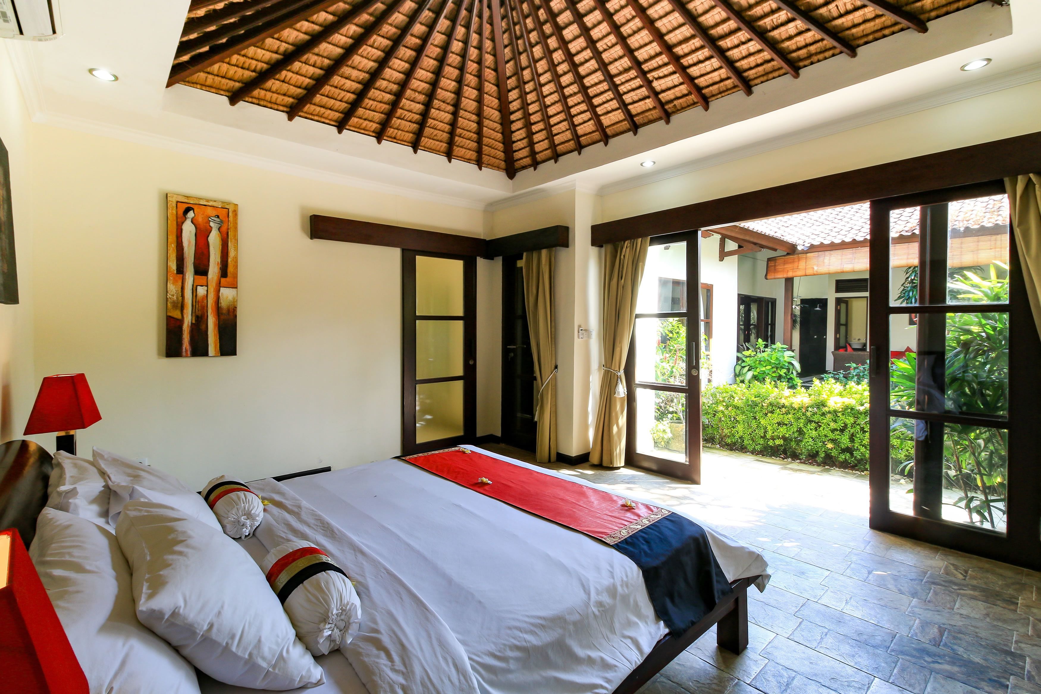 Quarto de hotel com uma cama, um quadro, um abajur vermelho e vista do jardim pela porta.