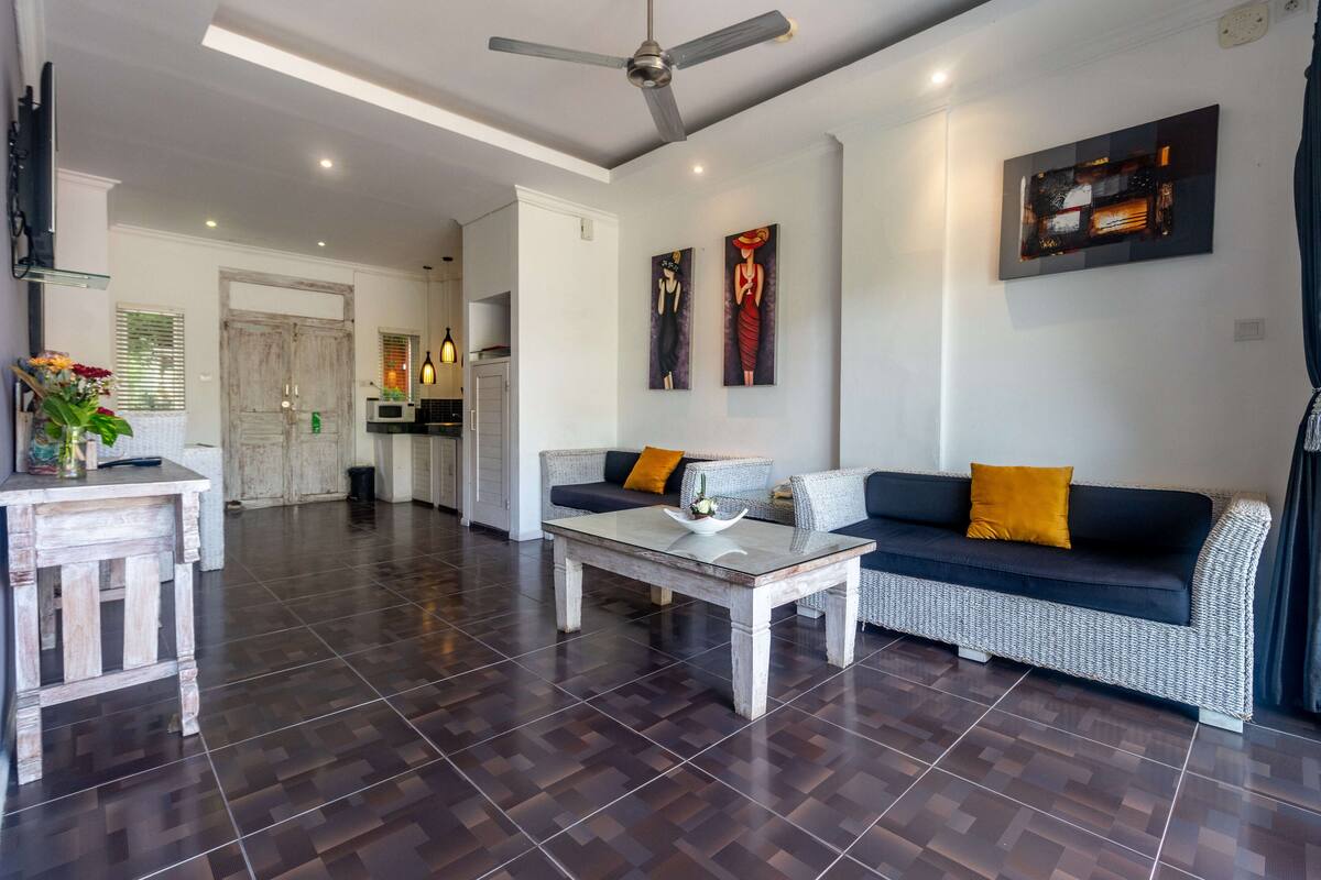 A spacious living area with a sofa, coffee table, and a dining table. There is a ceiling fan, artwork on the walls, and a kitchen in the background.
