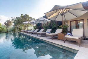 Zona junto a la piscina con sillones de descanso y sombrillas, rodeada de vegetación.