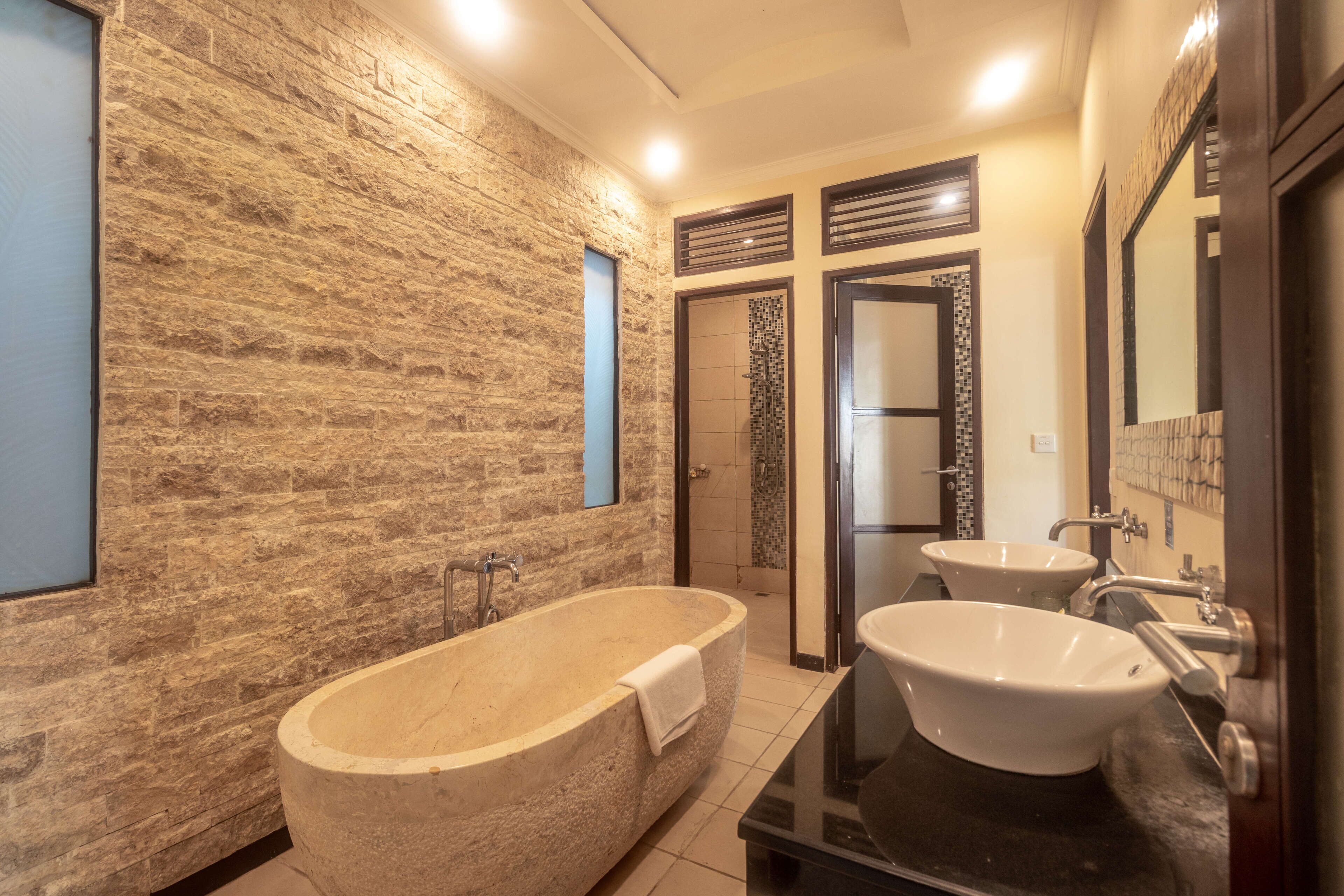 A modern bathroom with a stone wall, a freestanding bathtub, two sinks, and a large mirror.