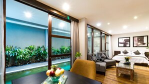 Une chambre d’hôtel moderne avec une grande fenêtre, une table  à manger avec des fruits, un canapé, un lit avec des oreillers et une petite table avec une plante.