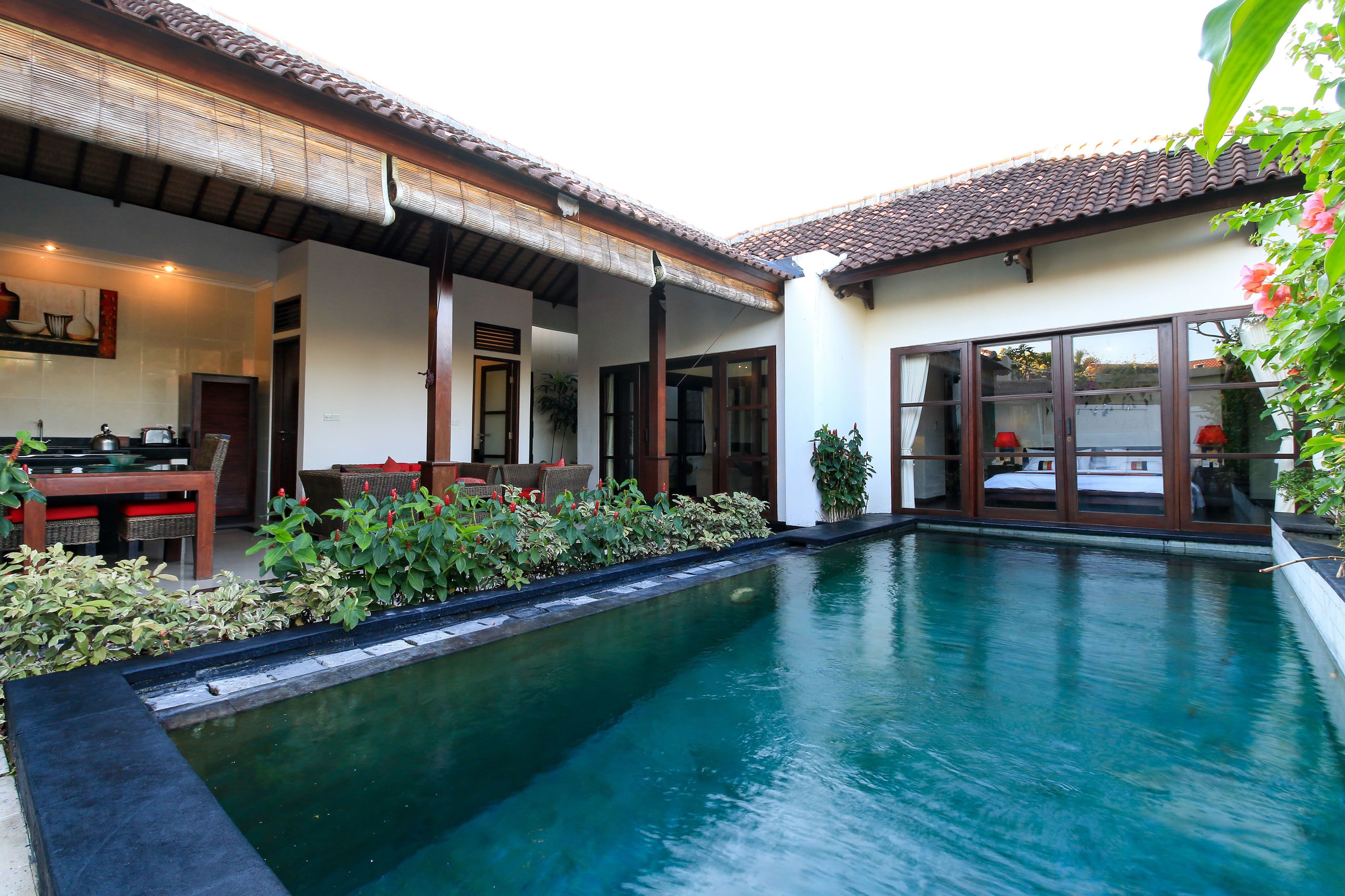 A swimming pool with a tiled deck, a dining area with a table and chairs, and a covered outdoor seating area with a view of the pool.