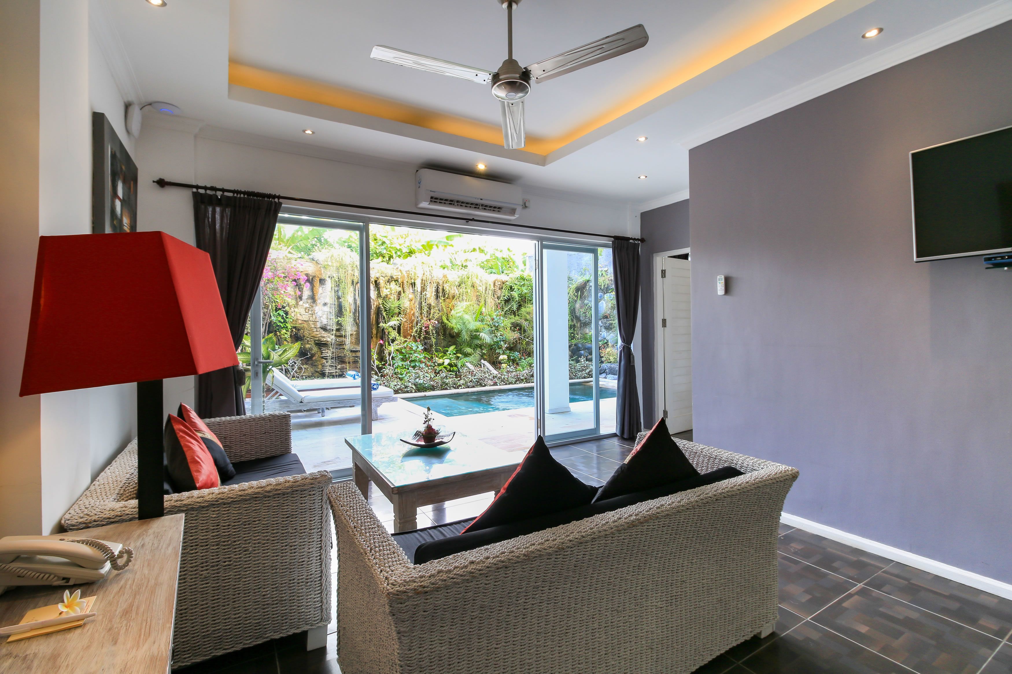 Una sala de estar moderna con muebles de mimbre, una lámpara roja y vistas a una piscina y jardín.