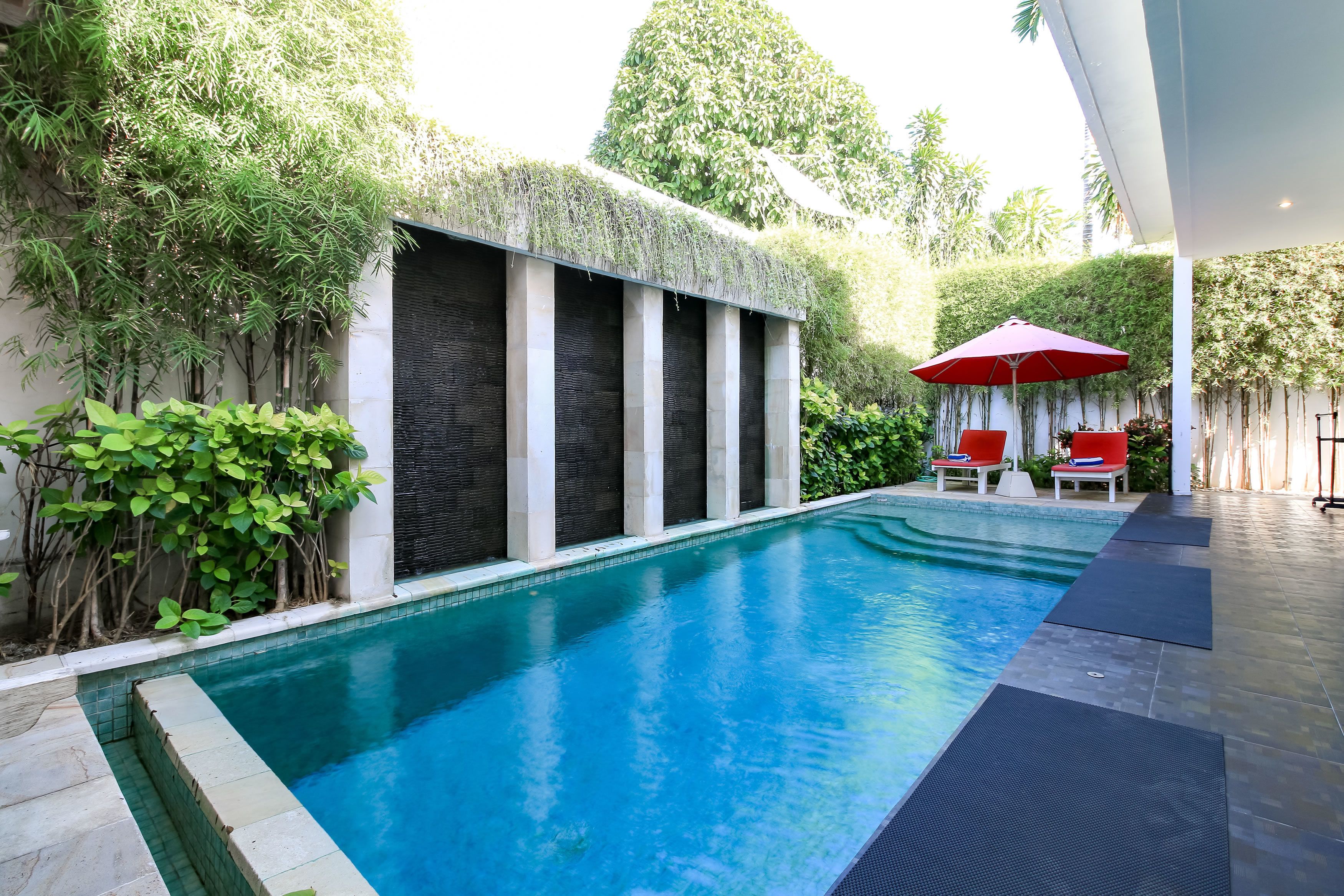 A swimming pool with a modern design, surrounded by lush greenery and a covered seating area with red umbrellas.