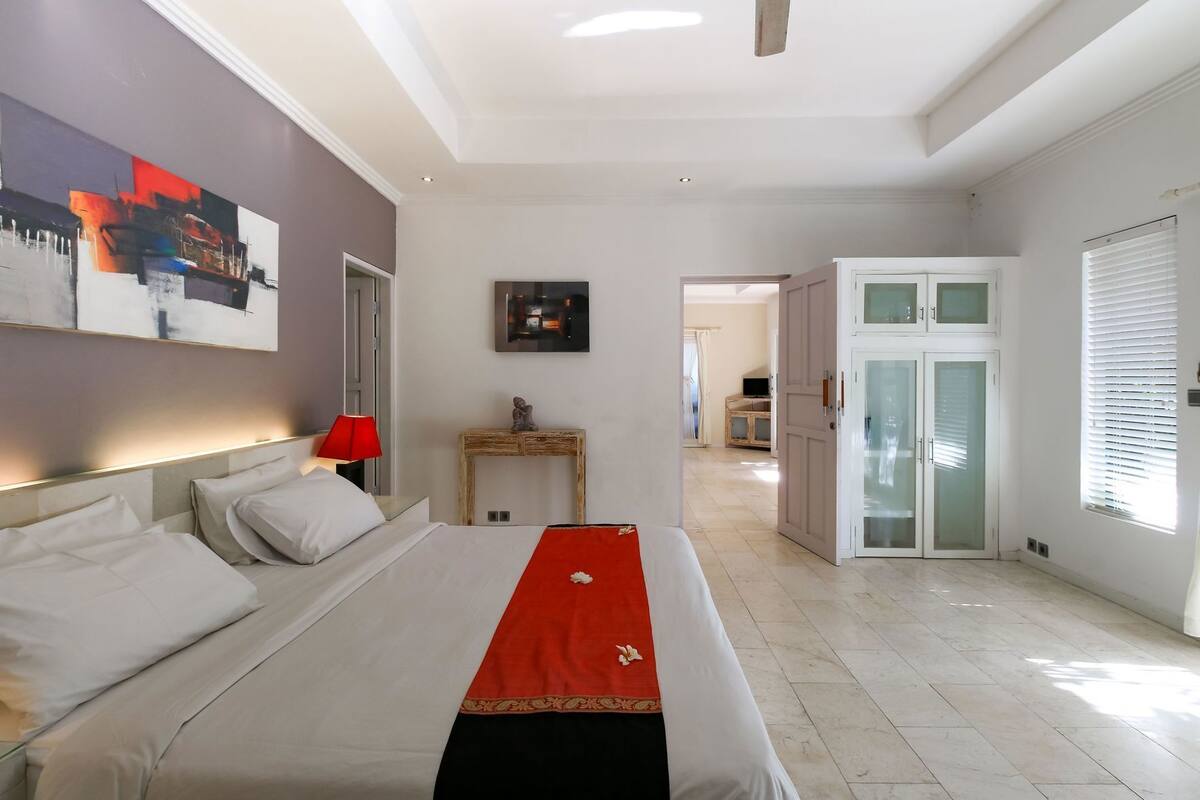 A hotel room with a bed, a red throw, a painting, a lamp, and a view of the hallway.