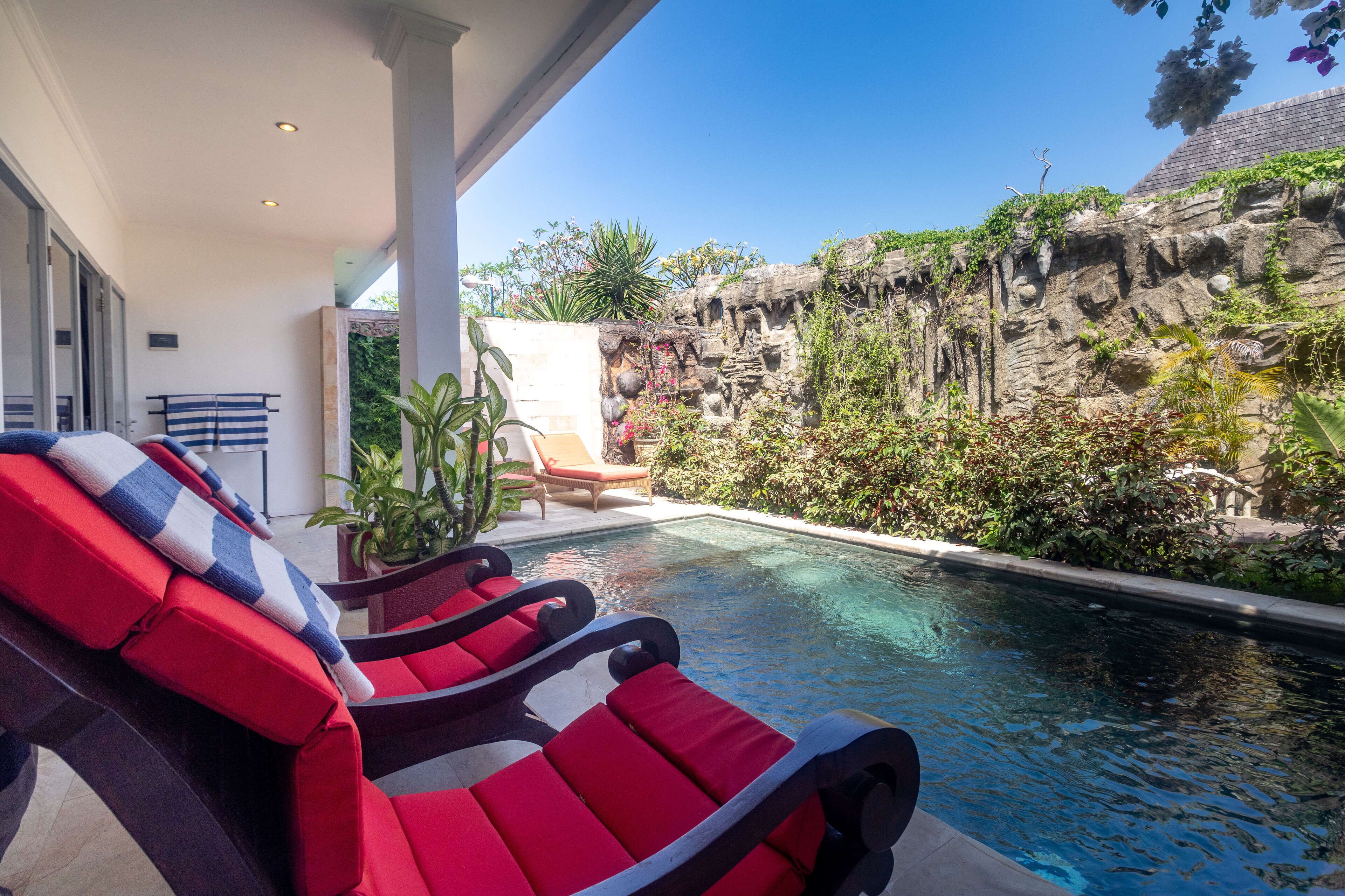 A pool area with red lounge chairs, a small table, and a potted plant.