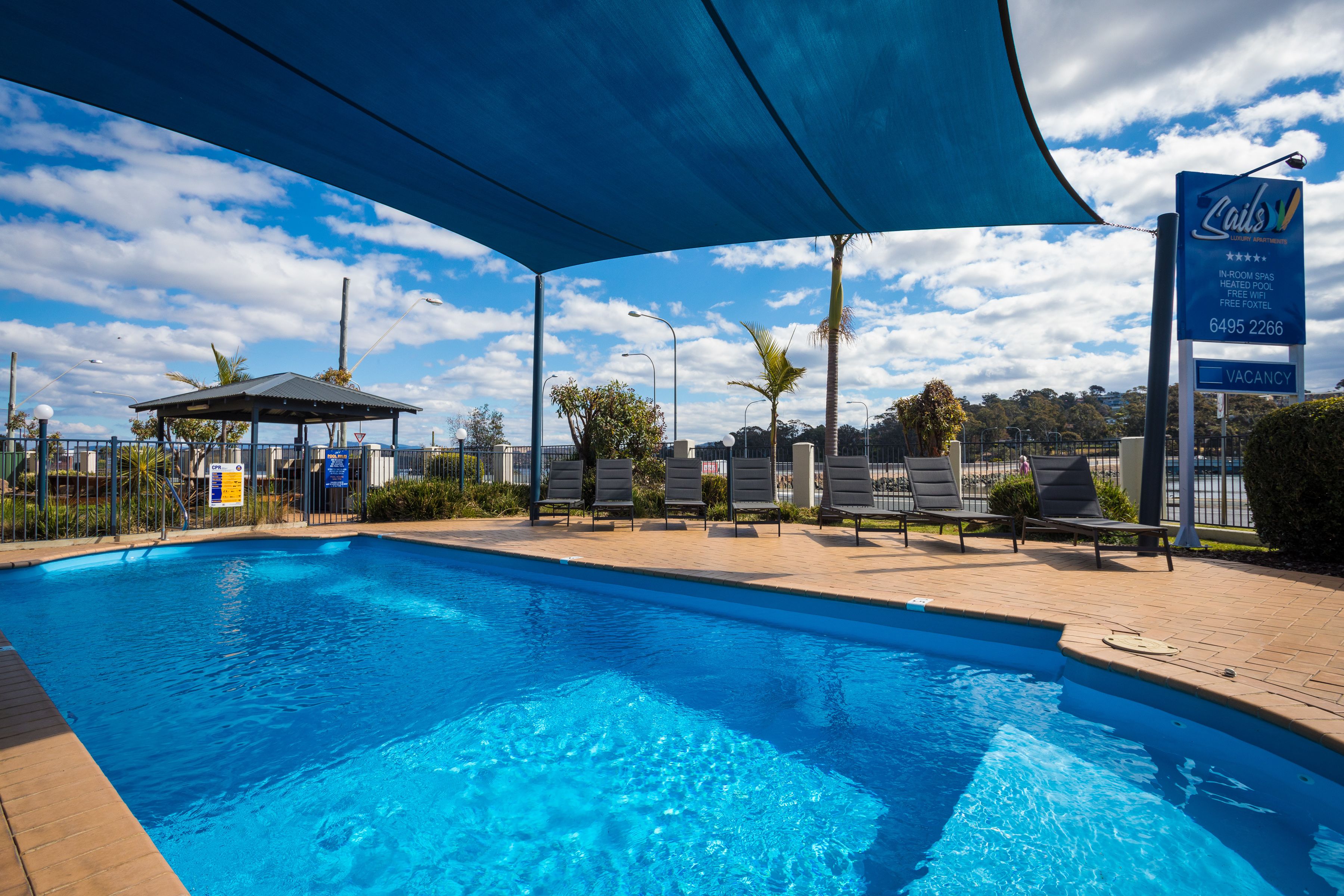 A swimming pool with lounge chairs and a shaded area, a signboard for a hotel, and a clear blue sky.