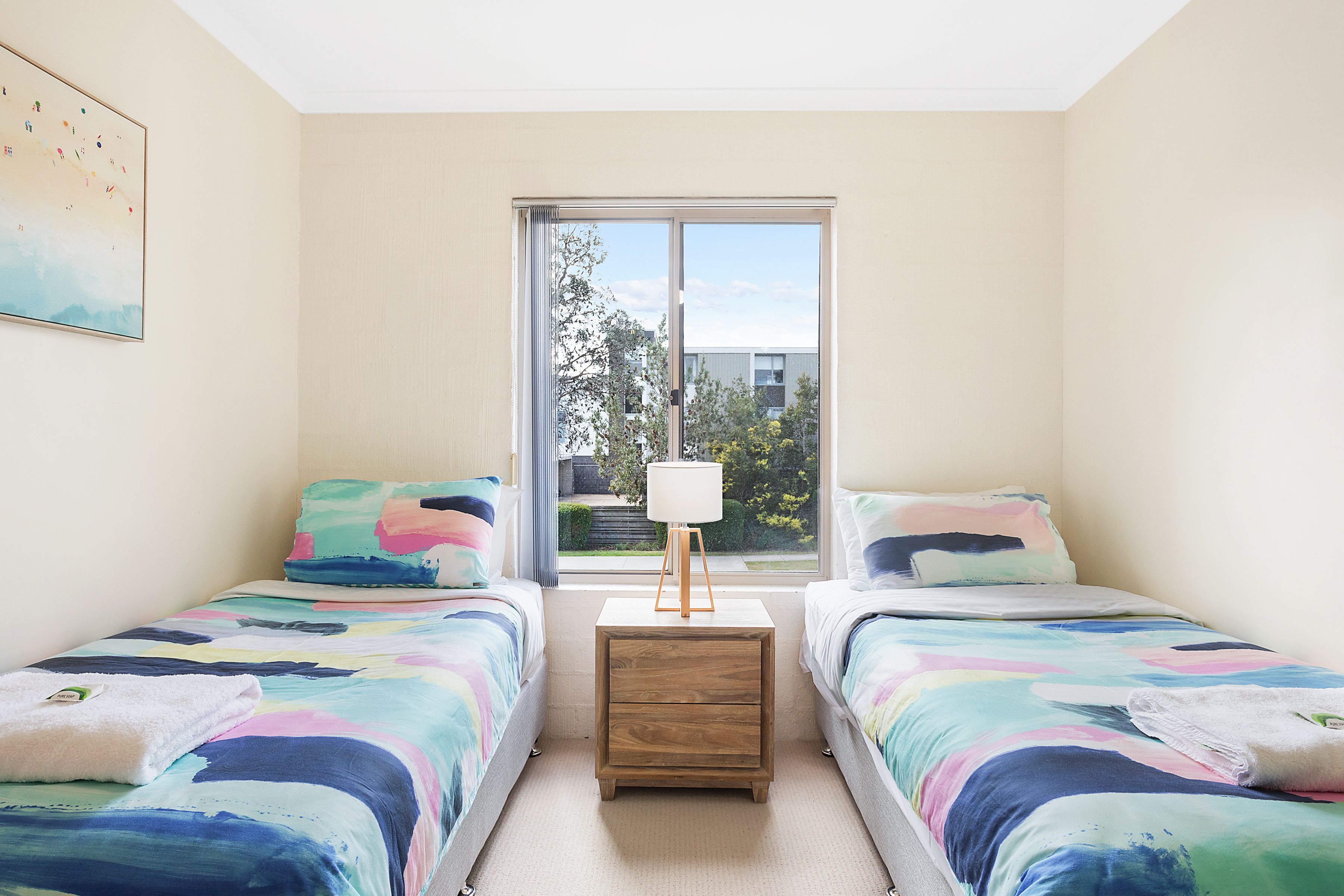 Una camera da letto con due letti, un comodino con una lampada e una finestra che si affaccia su edifici e alberi.
