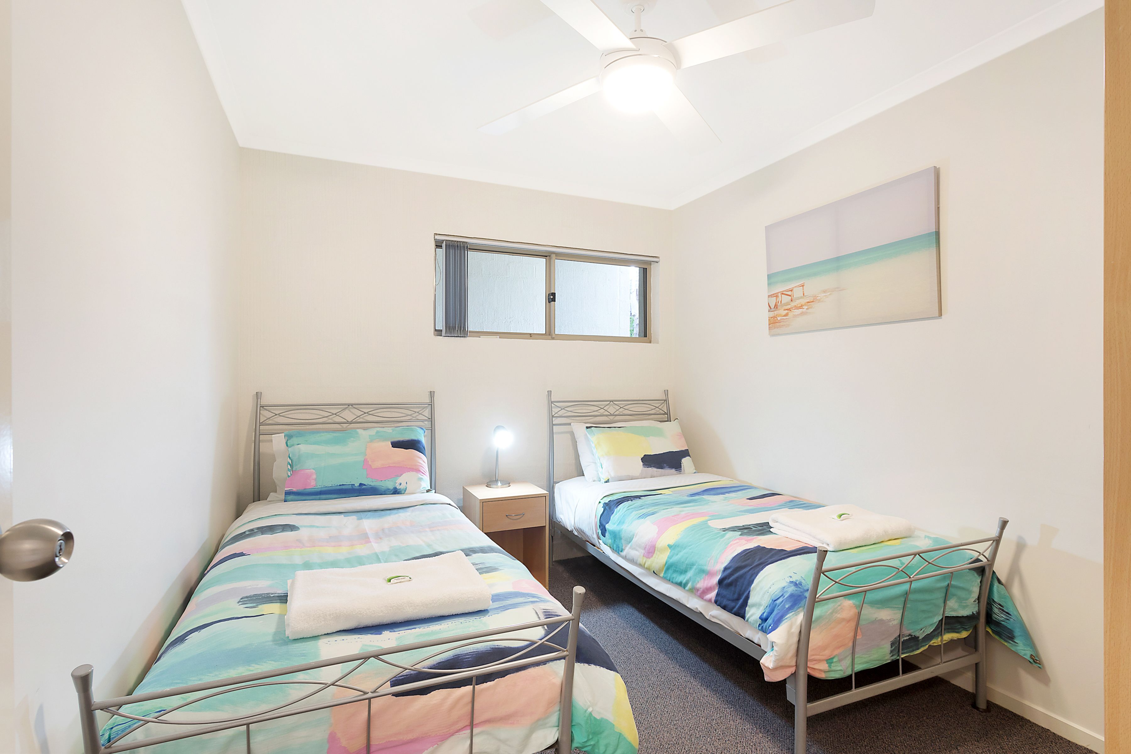 Un dormitorio pequeño y bien iluminado con dos camas individuales, un ventilador de techo, una ventana y un cuadro en la pared.