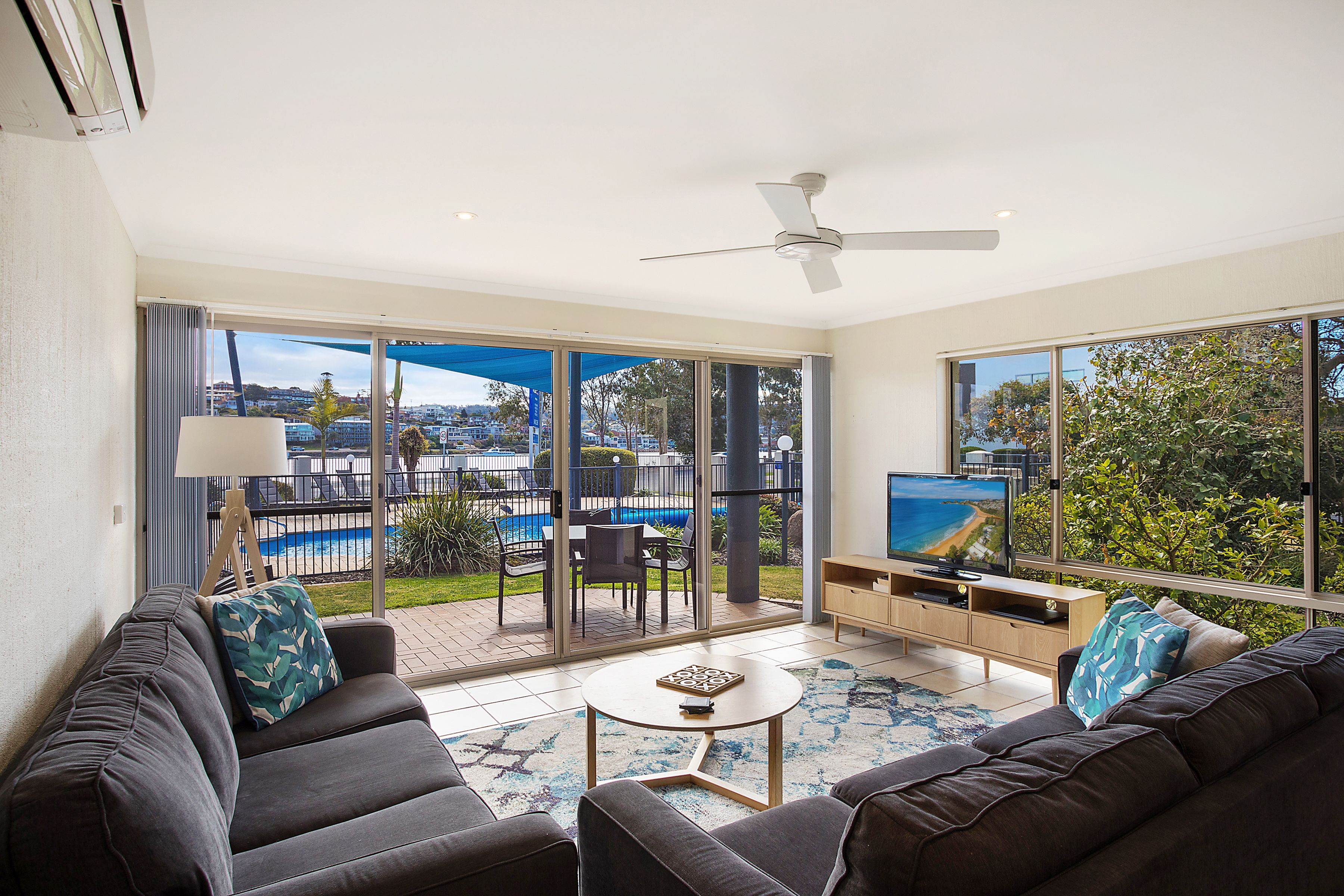 A modern living room with a large TV, comfortable seating, and a view of a pool and greenery.
