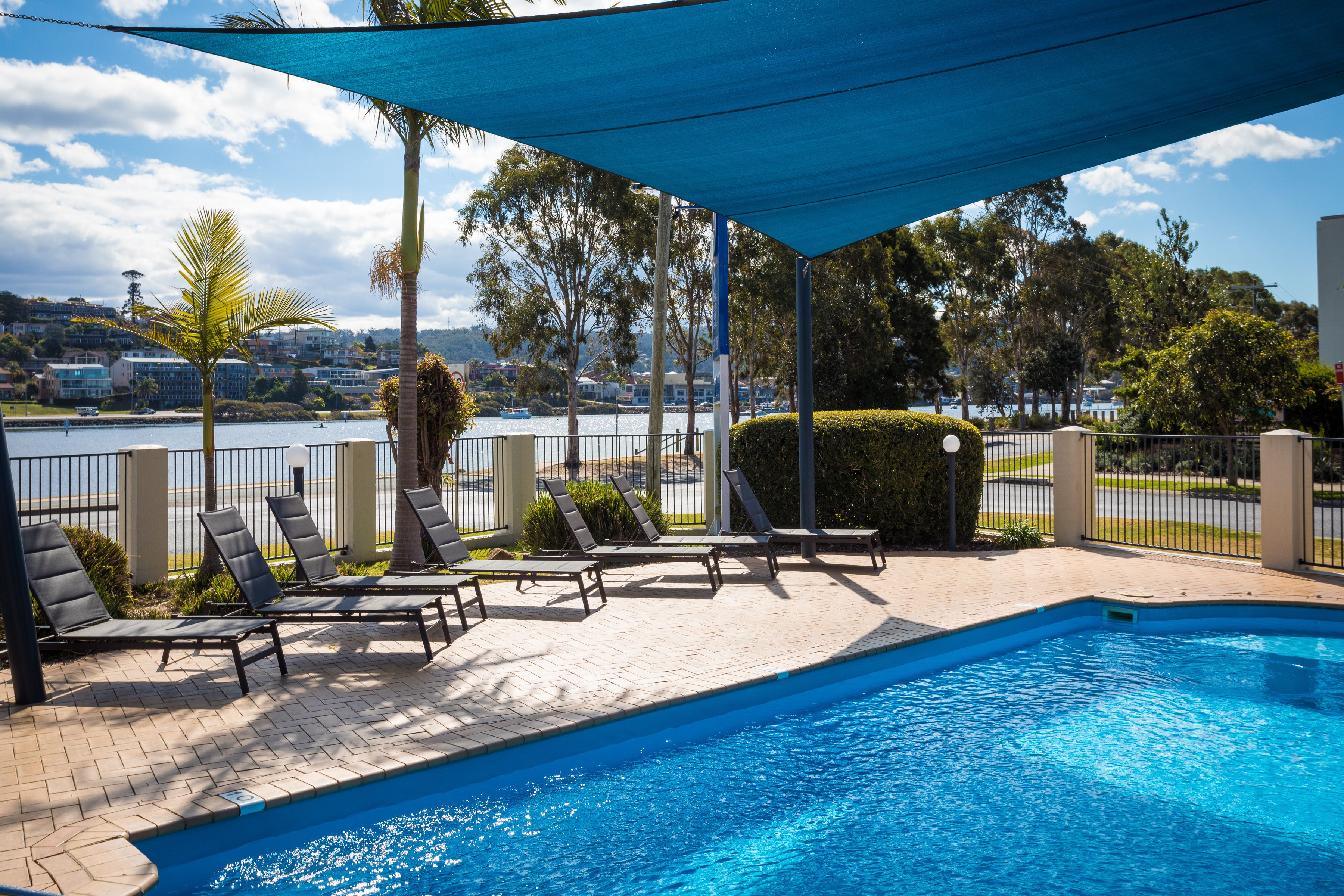 Una piscina con sillas de descanso y una zona sombreada, rodeada por una cerca y follaje.