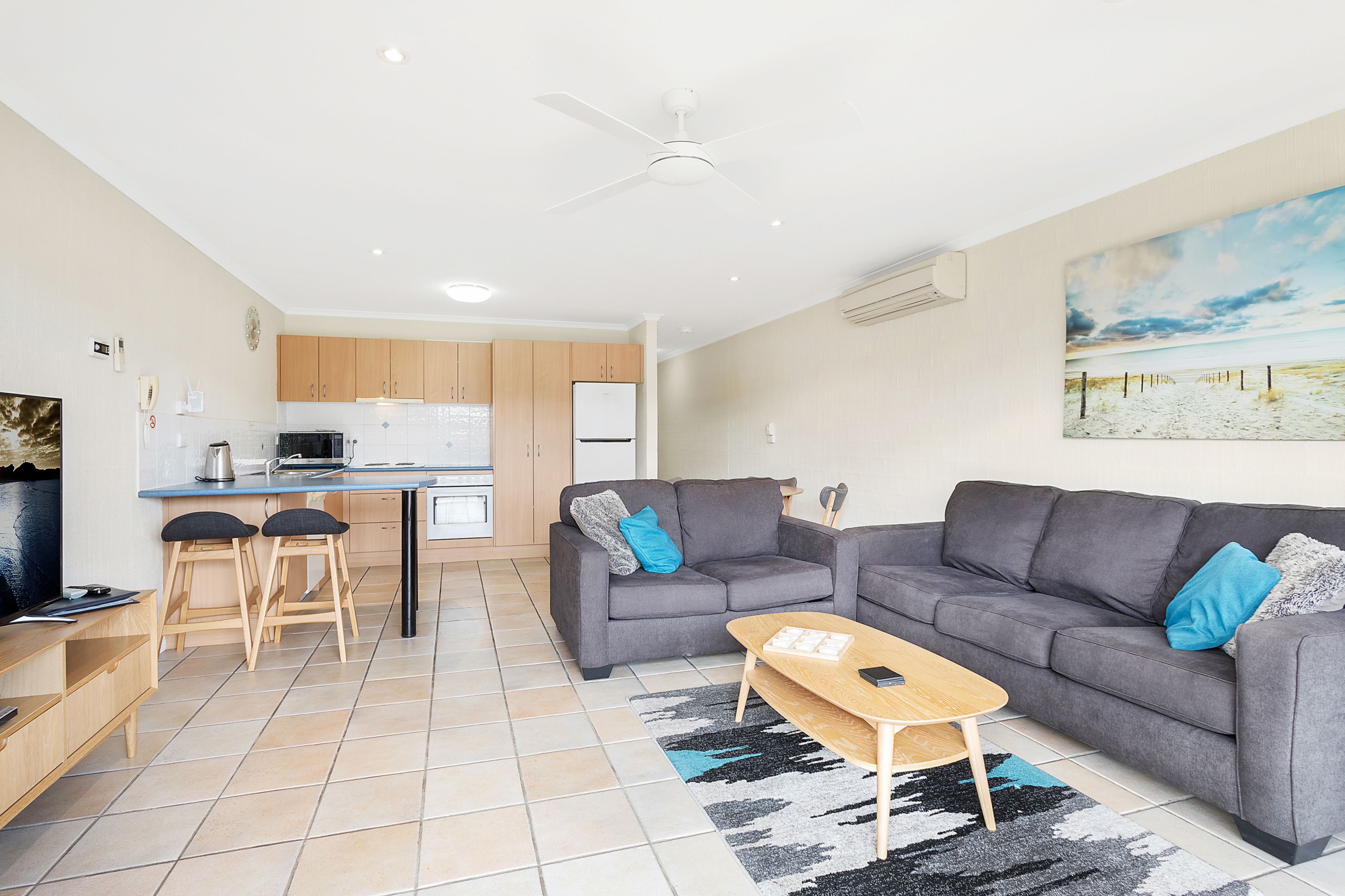 A modern living room with a grey sofa, a coffee table, and a TV. There is a kitchen area with wooden cabinets and a white fridge.
