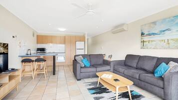 A modern living room with a grey sofa, a coffee table, and a TV. There is a kitchen area with wooden cabinets and a white fridge.
