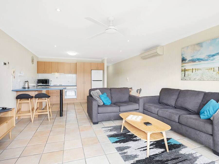 A modern living room with a grey sofa, a coffee table, and a TV. There is a kitchen area with wooden cabinets and a white fridge.