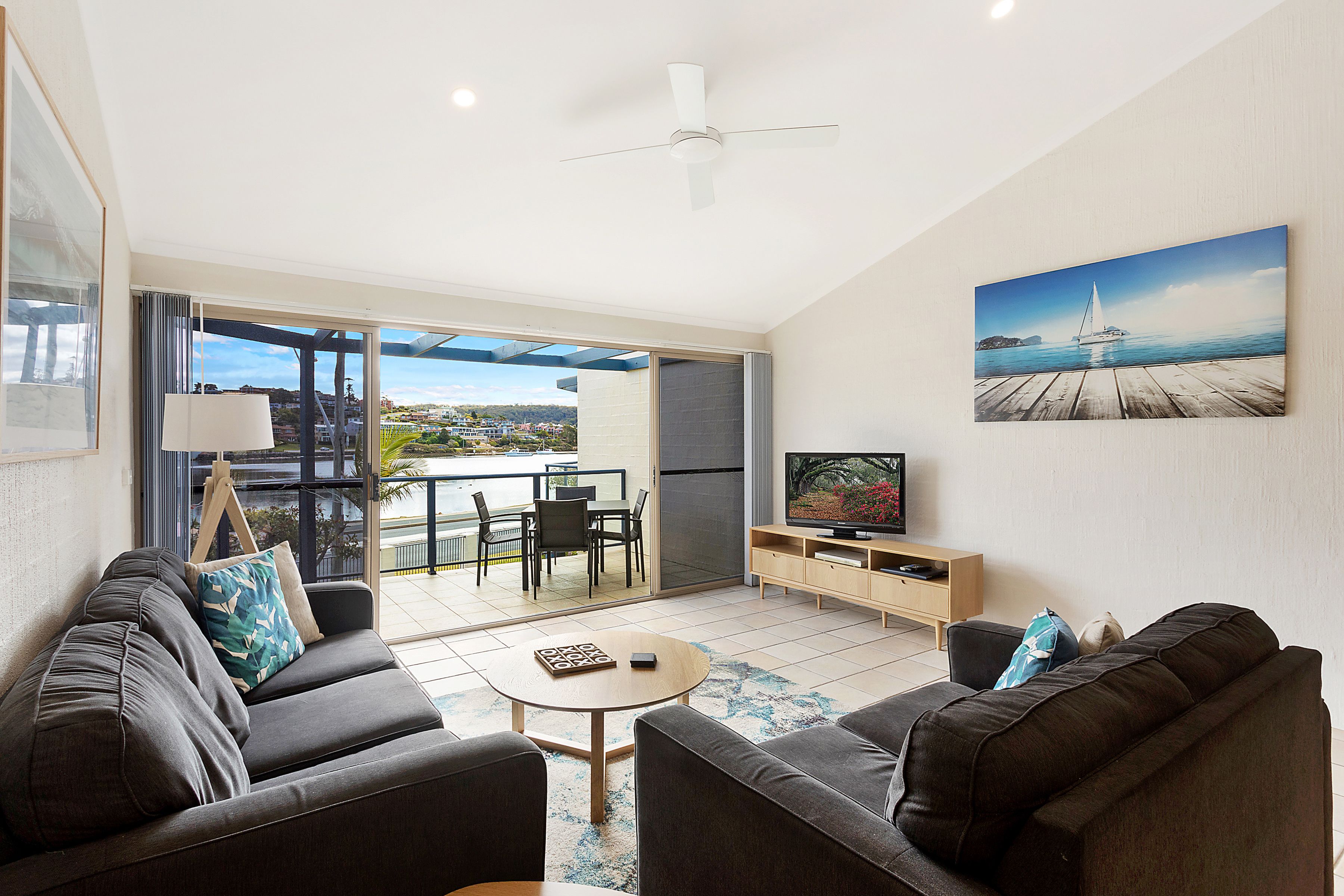 Un salon moderne avec un canapé, une table basse et un téléviseur, un balcon avec vue sur la mer et un ventilateur de plafond.
