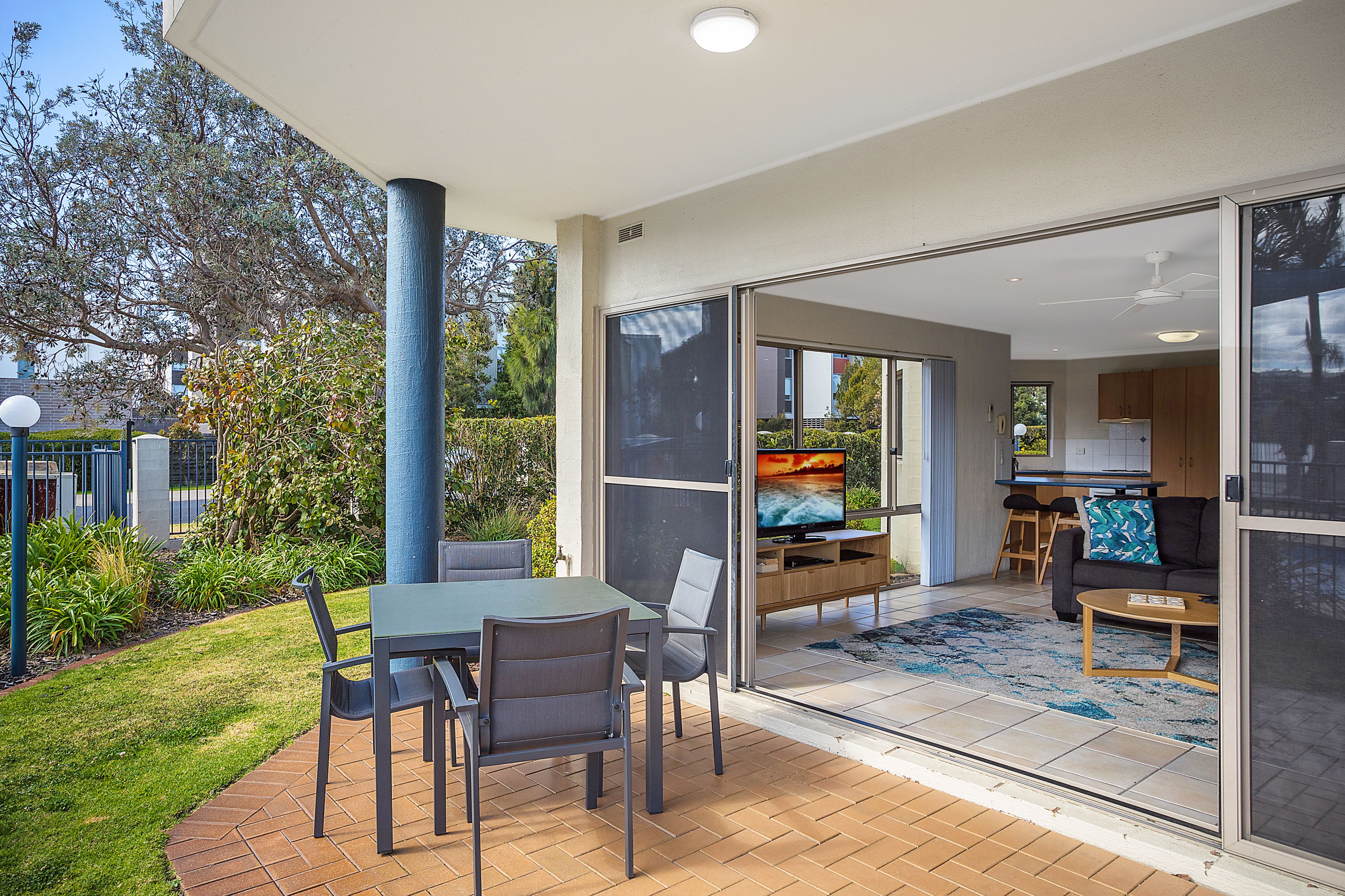 A modern outdoor patio area with a dining table and chairs, a TV, and a kitchen visible through glass doors.
