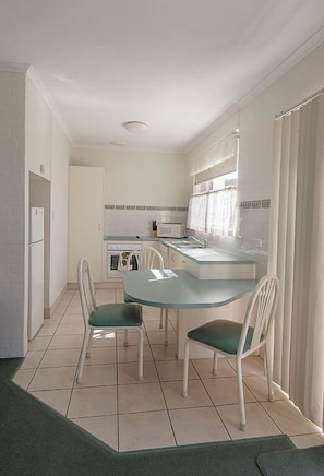 A compact kitchen with a dining area, white cabinetry, a small oven, and a window with curtains. - Merimbula Beach Apartments (Merimbula)