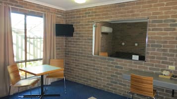 Une pièce avec un mur en briques, un téléviseur à écran plat fixé au-dessus d’un miroir, une petite table avec une chaise, et une deuxième chaise avec un bureau.