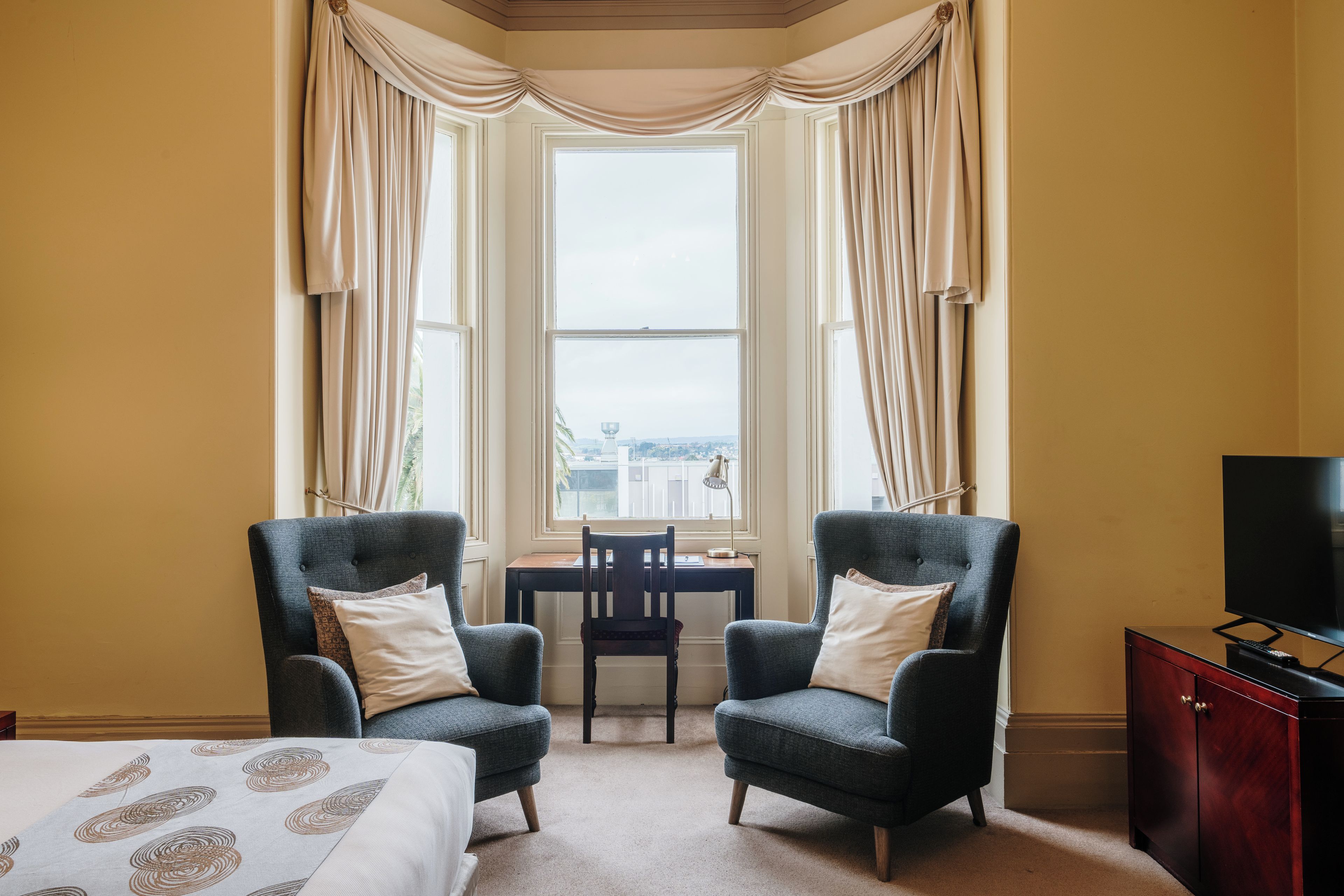 A hotel room with two armchairs, a bed, a desk, and a television.