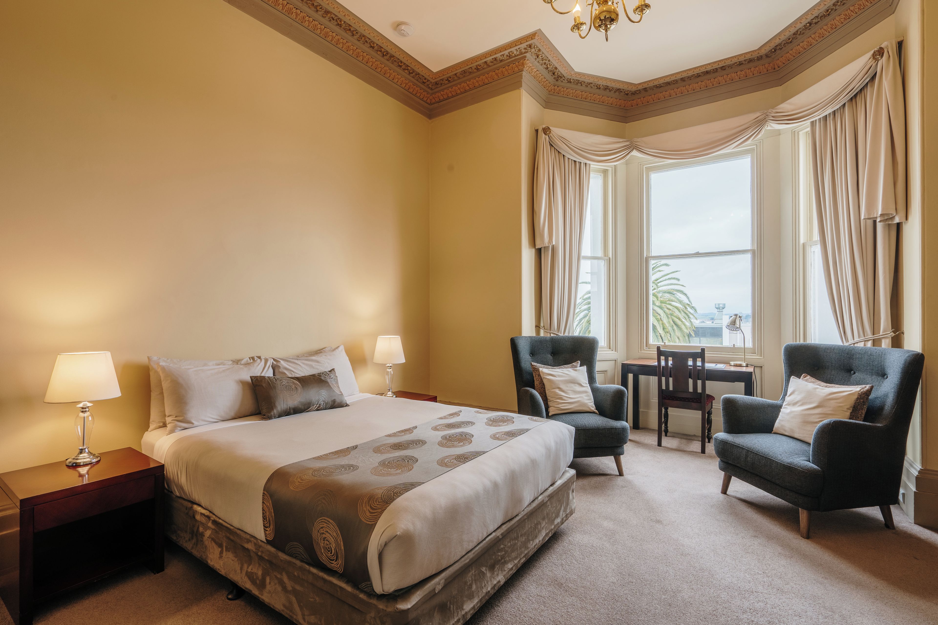 A hotel room with a large bed, two armchairs, a desk, and a view of the outdoors through the windows.
