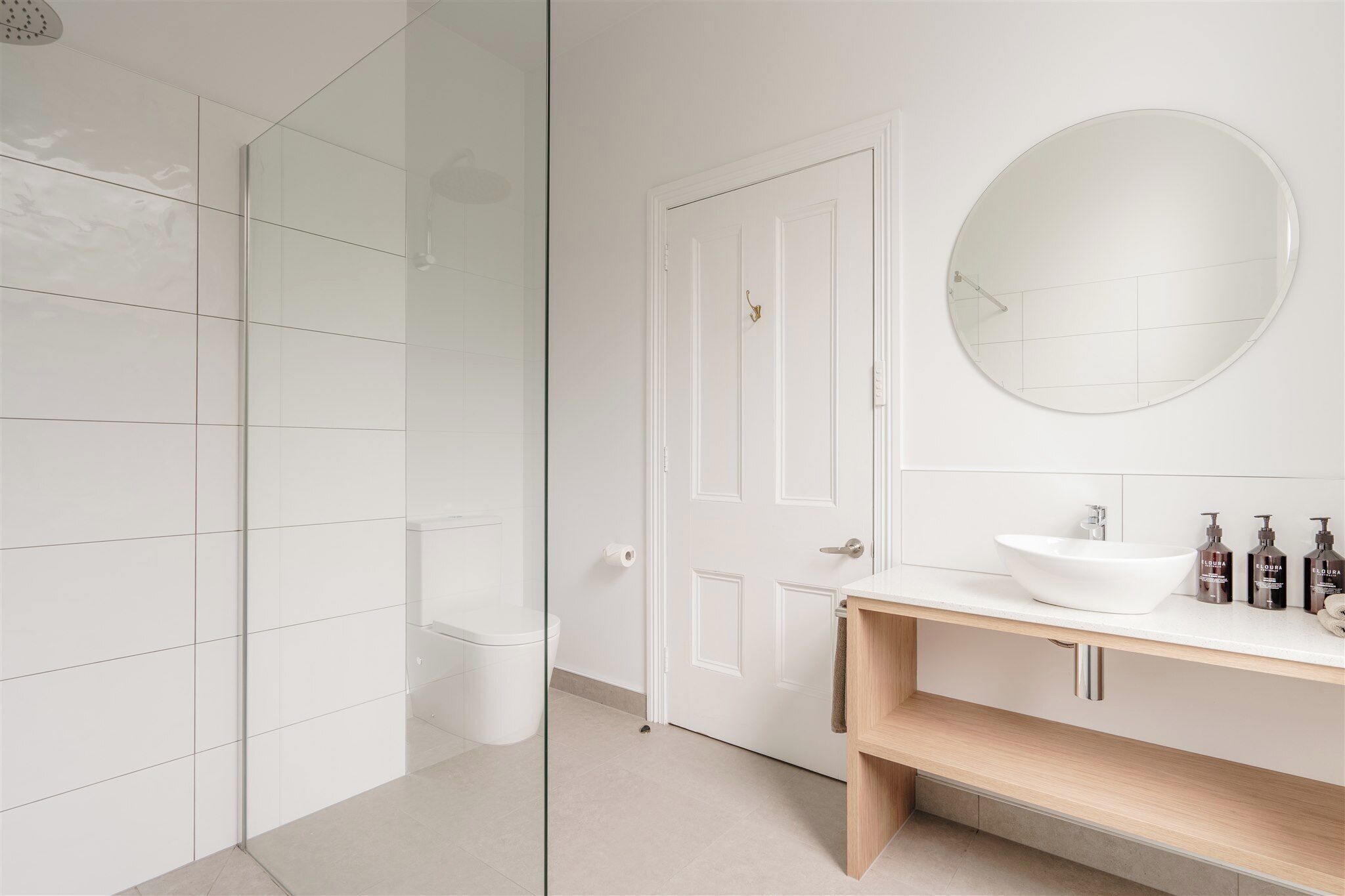 Un bagno moderno con cabina doccia in vetro, water bianco, specchio rotondo e mobile lavabo in legno con lavandino bianco.