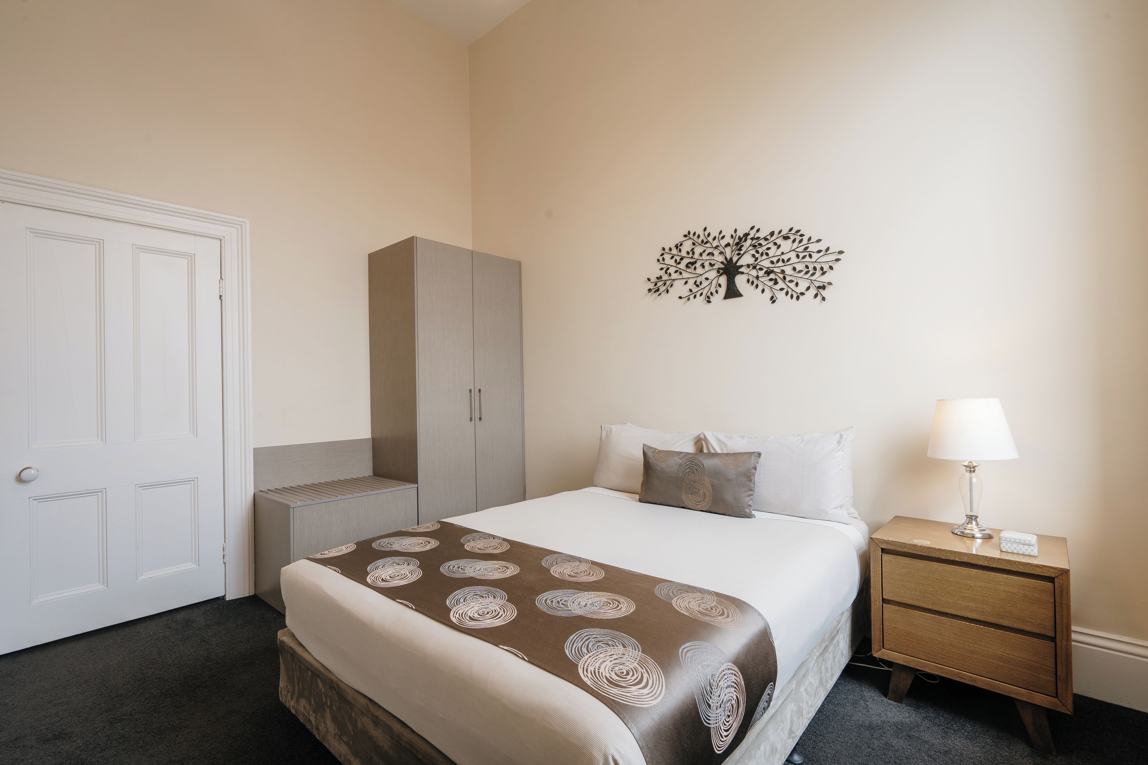 Ein Hotelzimmer mit Bett, Nachttisch, Lampe, Kleiderschrank und Wanddeko.