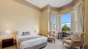 A hotel room with a large bed, two chairs, a desk, and a view of palm trees through the window. - Waratah on York (Launceston)