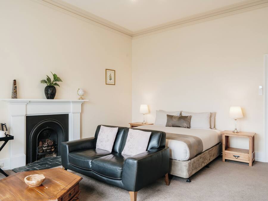 Une chambre d’hôtel avec un canapé en cuir noir, une table basse en bois, un lit avec deux oreillers, une table de chevet avec une lampe et une cheminée avec une manteau de cheminée.