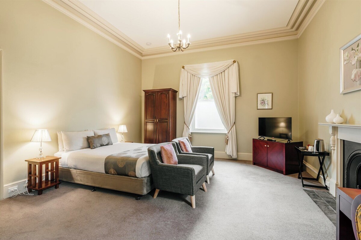 A bedroom with a bed, two armchairs, a wooden cabinet, a television, and a fireplace.