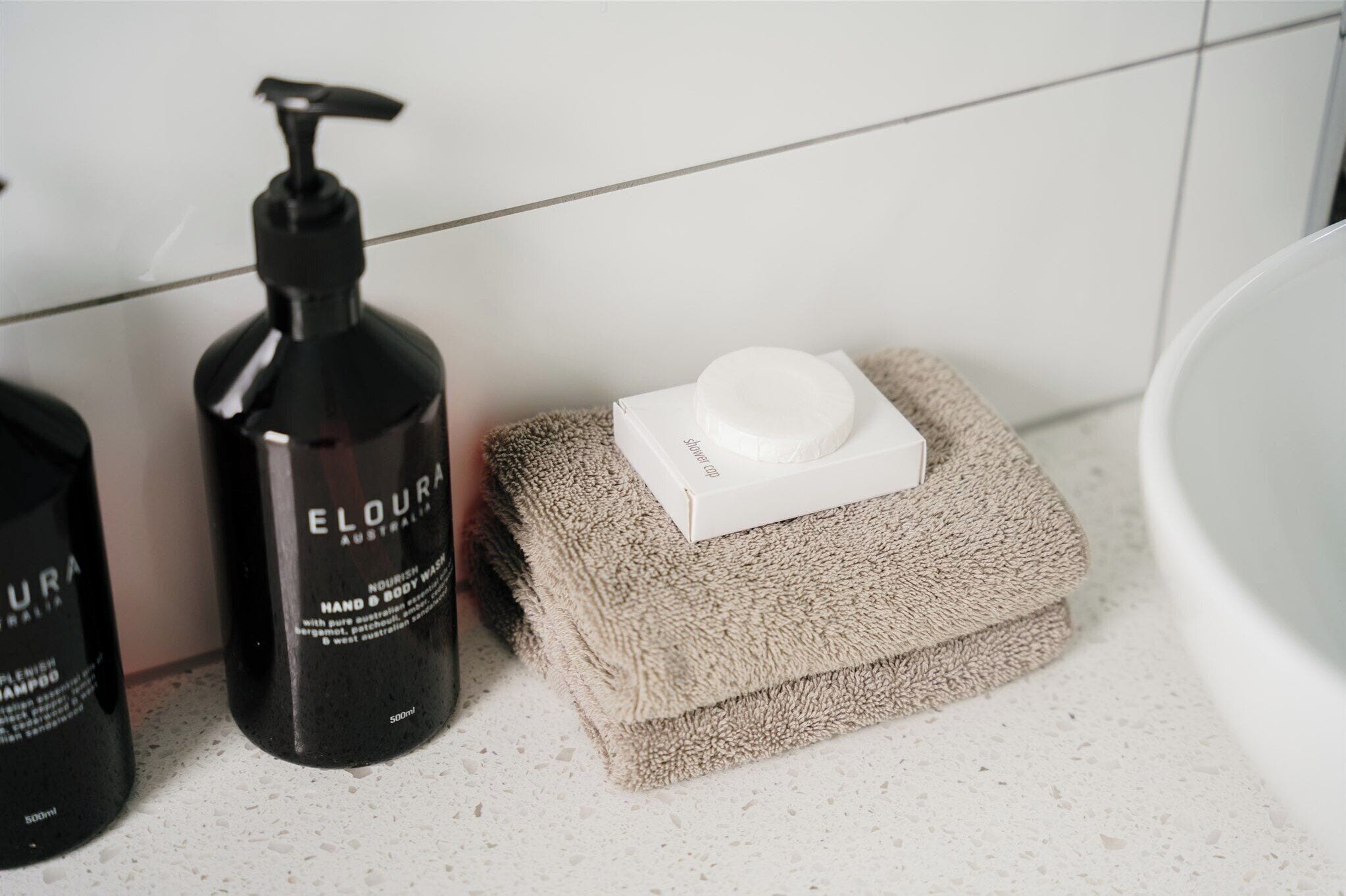 Ein Badezimmer mit einem weißen Waschbecken, einer schwarzen Flasche Eloura Hand- und Körperpflege sowie einem beigefarbenen Handtuch auf einer weißen Arbeitsplatte.