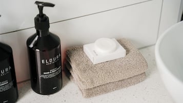 Ein Badezimmer mit einem weißen Waschbecken, einer schwarzen Flasche Eloura Hand- und Körperpflege sowie einem beigefarbenen Handtuch auf einer weißen Arbeitsplatte.