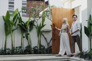 A couple standing in front of a wooden door, with a garden and a building in the background. - Gracia Bali Villas & Apartment (Seminyak)