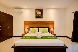 A hotel room with a large bed, wooden headboard, two bedside lamps, a green bedspread, and a wall-mounted air conditioner.
