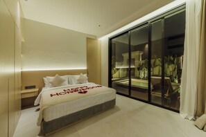 A hotel room with a large bed, a nightstand, a sliding glass door leading to a balcony with plants, and a view of the night sky. - Gracia Bali Villas & Apartment (Seminyak)