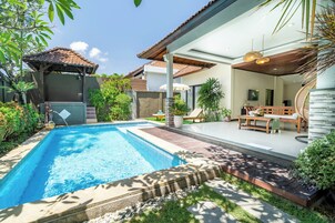 Una piscina con bordo piastrellato, una zona relax coperta con tavolo e sedie, e una scala che conduce a un livello superiore.