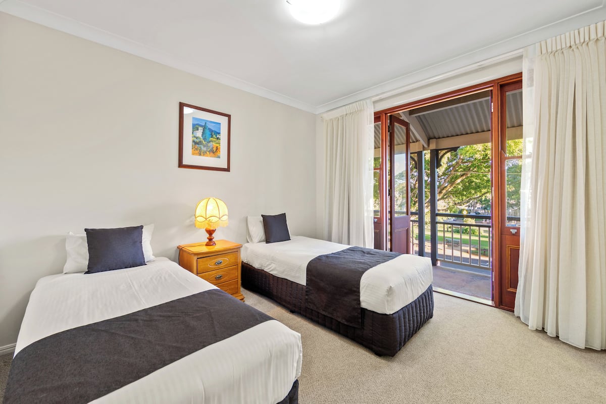 Une chambre d’hôtel avec deux lits, une table de chevet avec une lampe, un tableau encadré au mur et un balcon avec des rideaux.