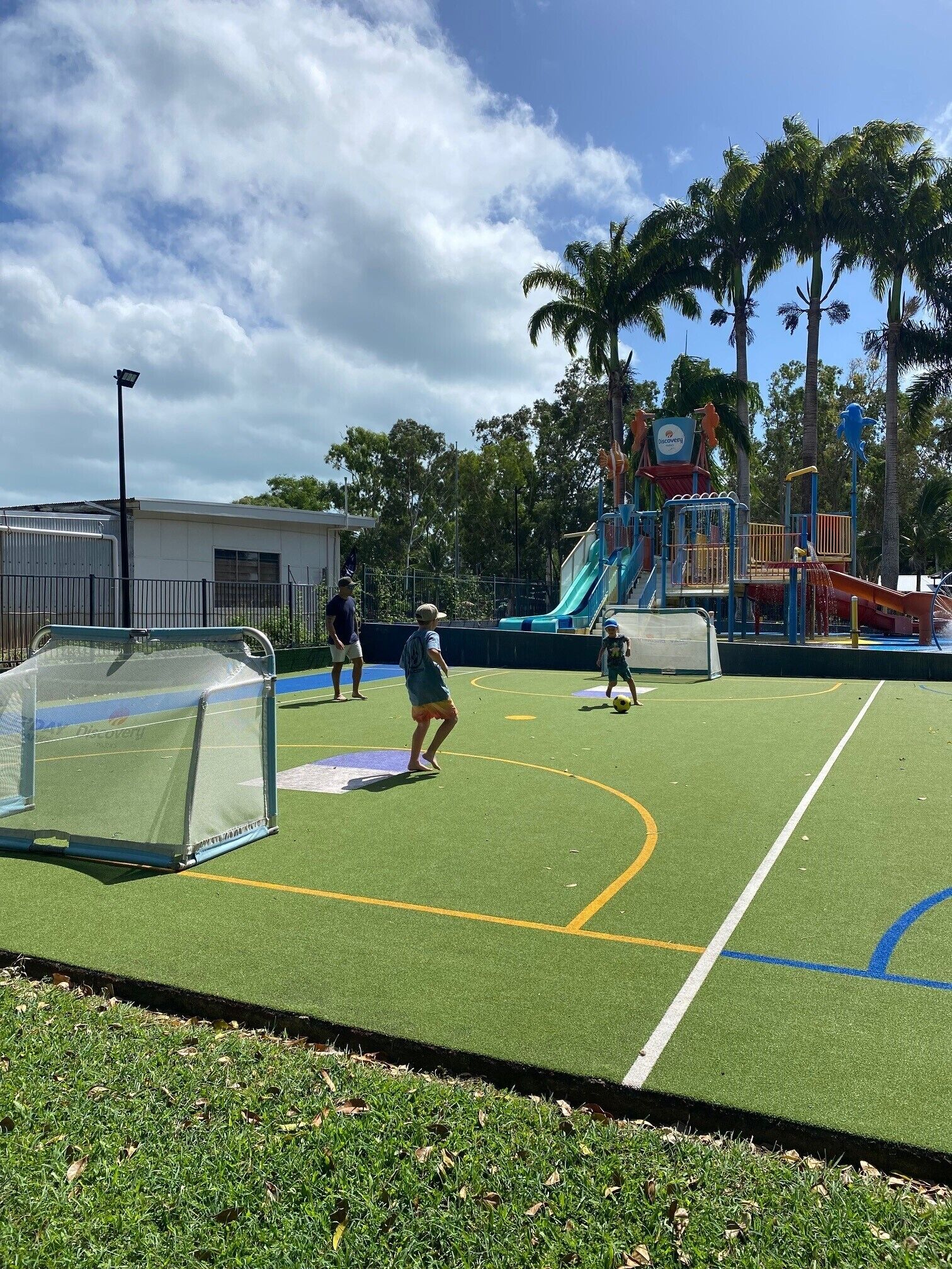 A sports court with a goalpost, a playground with slides, and palm trees in the background.