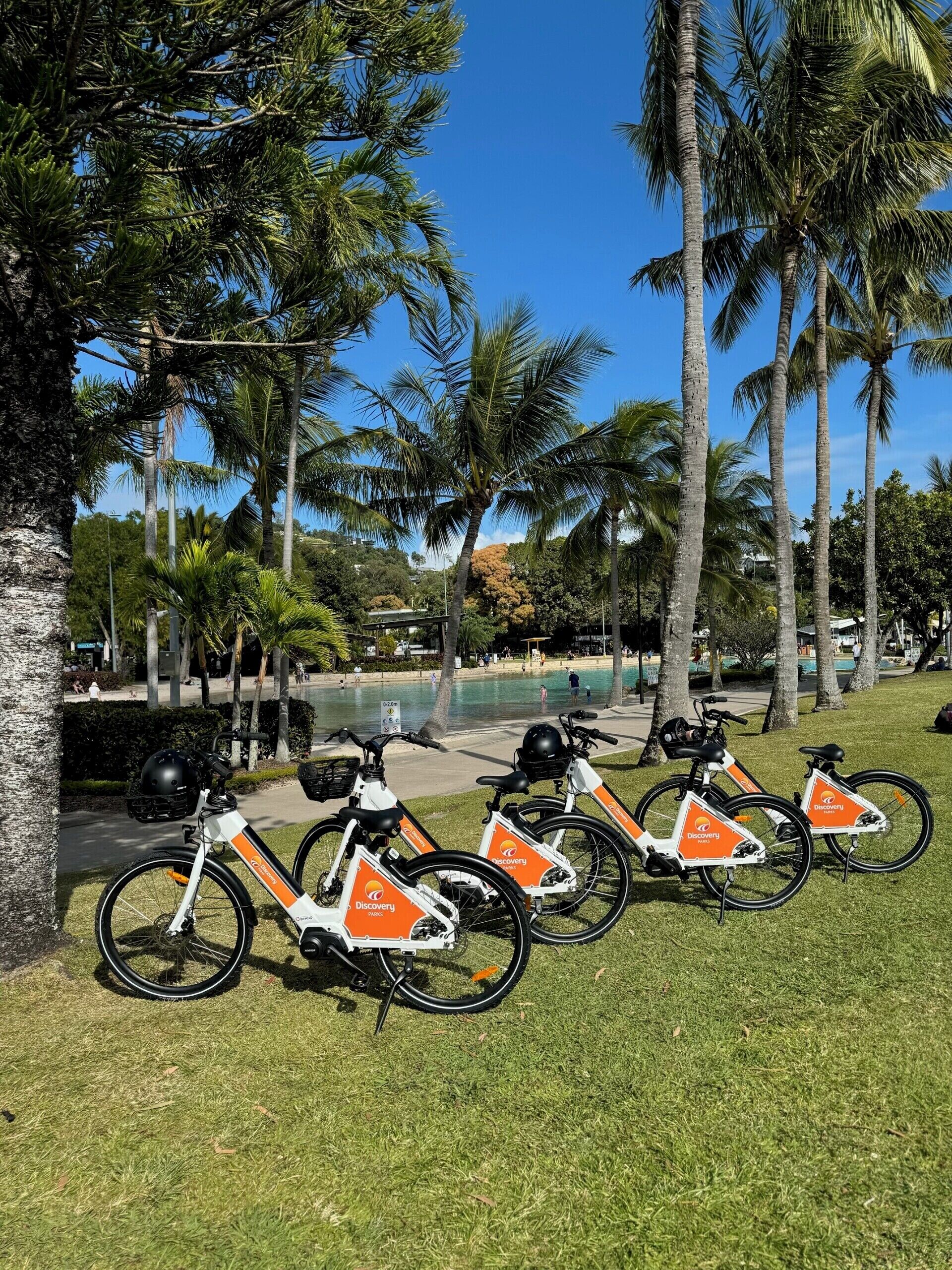 Una fila de bicicletas eléctricas con el logotipo naranja y blanco, estacionadas en un área verde con palmeras y una piscina al fondo.