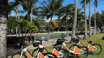 Une rangée de vélos électriques aux couleurs orange et blanche, garés sur une pelouse, avec des palmiers et une piscine en arrière-plan.