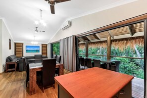 A modern living room with a dining area, large windows, ceiling fans, and a view of greenery. - Discovery Parks - Airlie Beach (Jubilee Pocket)