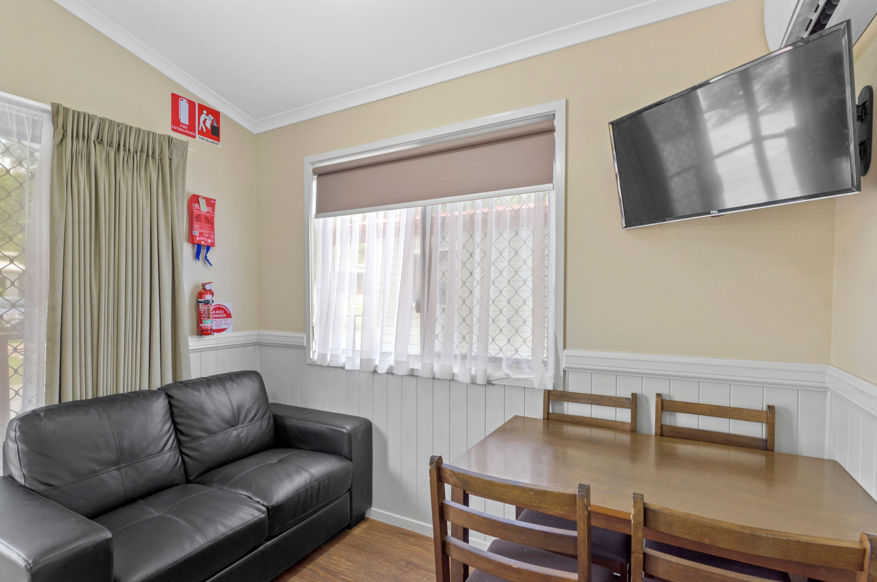 Una habitación con un sofá de cuero negro, una mesa de comedor de madera con cuatro sillas, un televisor montado en la pared y una ventana con cortinas blancas.