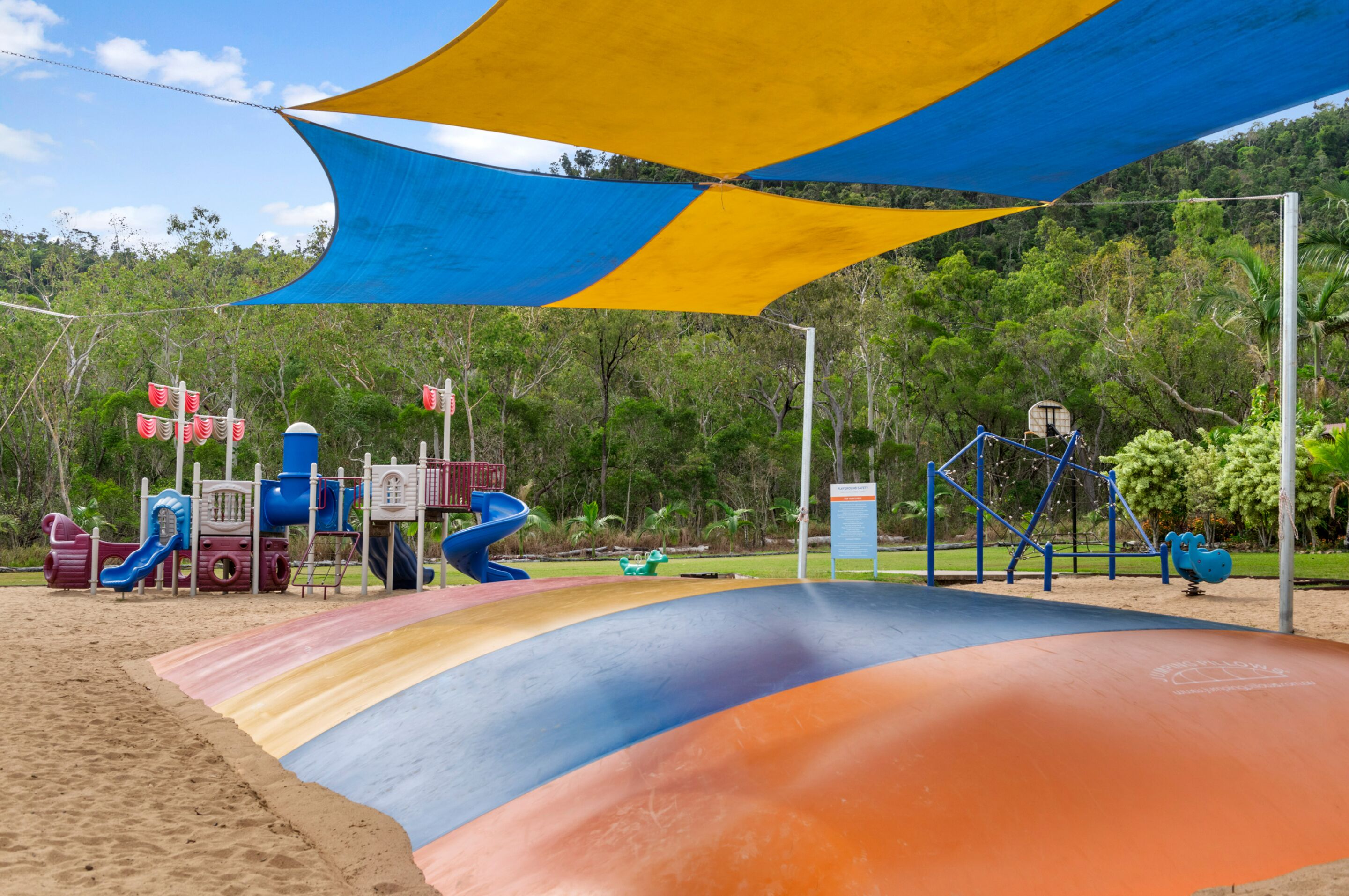 A playground with slides and climbing structures, surrounded by trees and a sandy area.