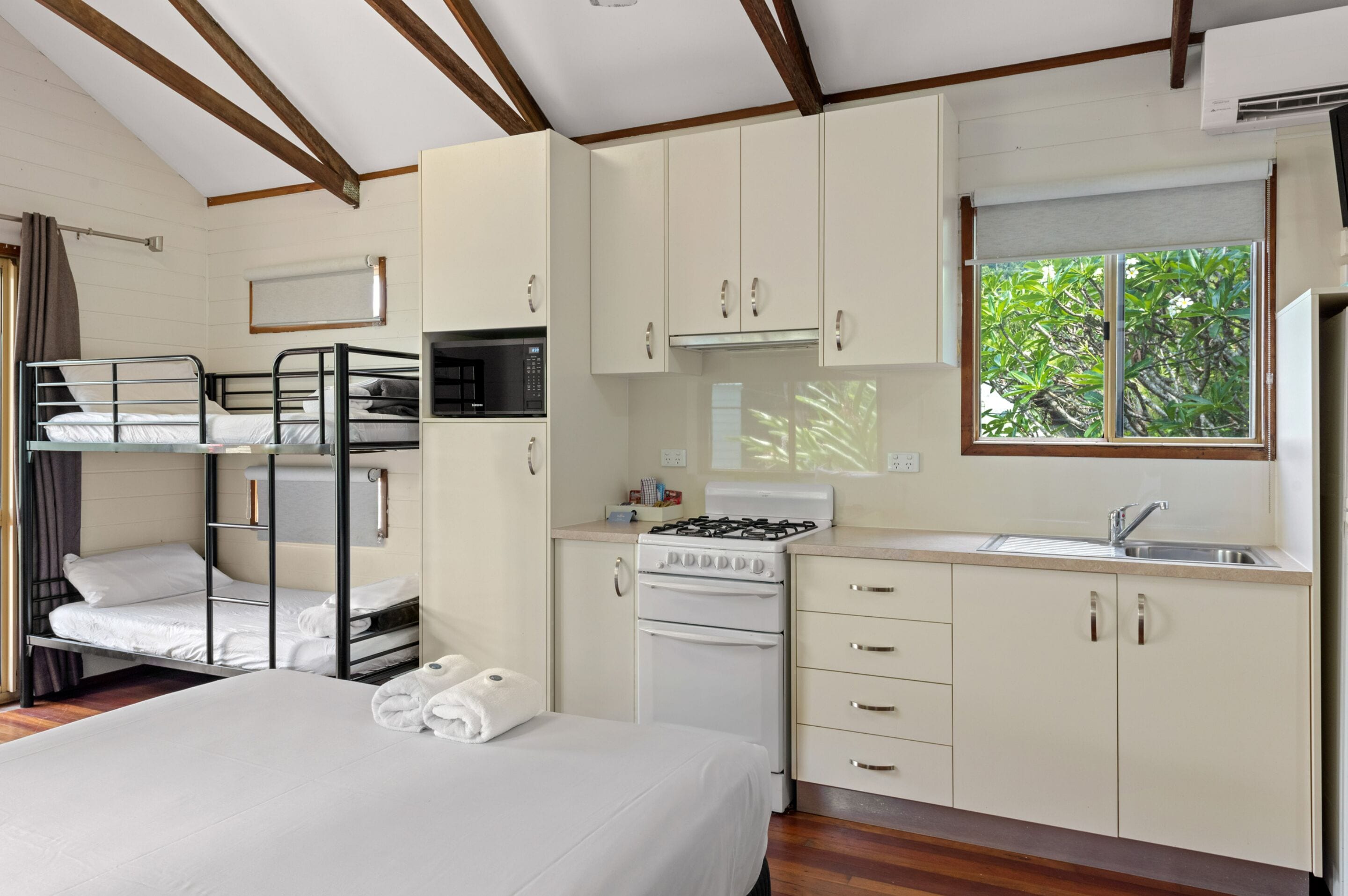 A compact room with bunk beds, a kitchenette with a stove and microwave, and a window with a view of greenery.