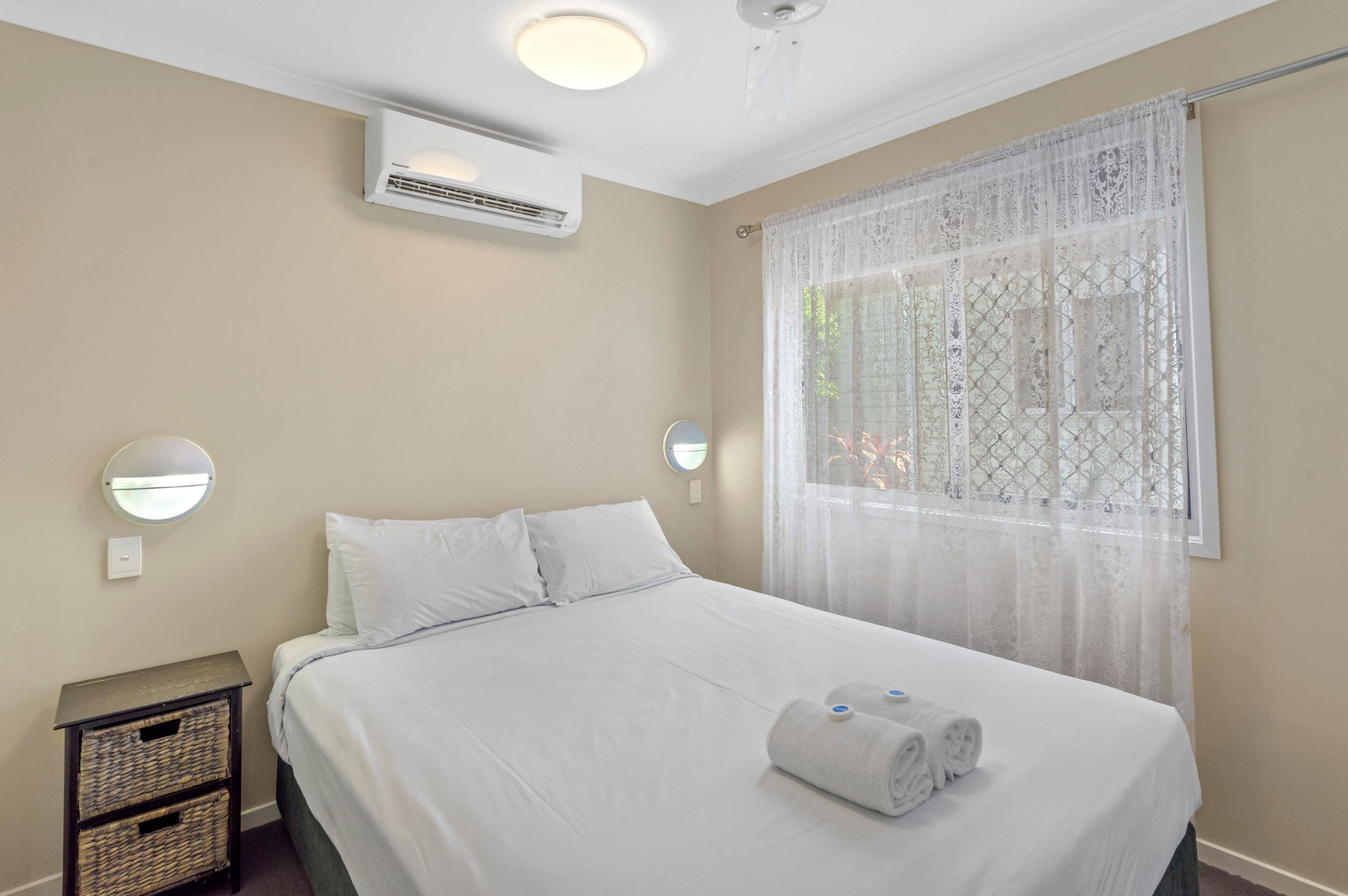 Una cama bien hecha con sábanas blancas, una mesita de noche con una cesta y una ventana con cortinas traslúcidas.