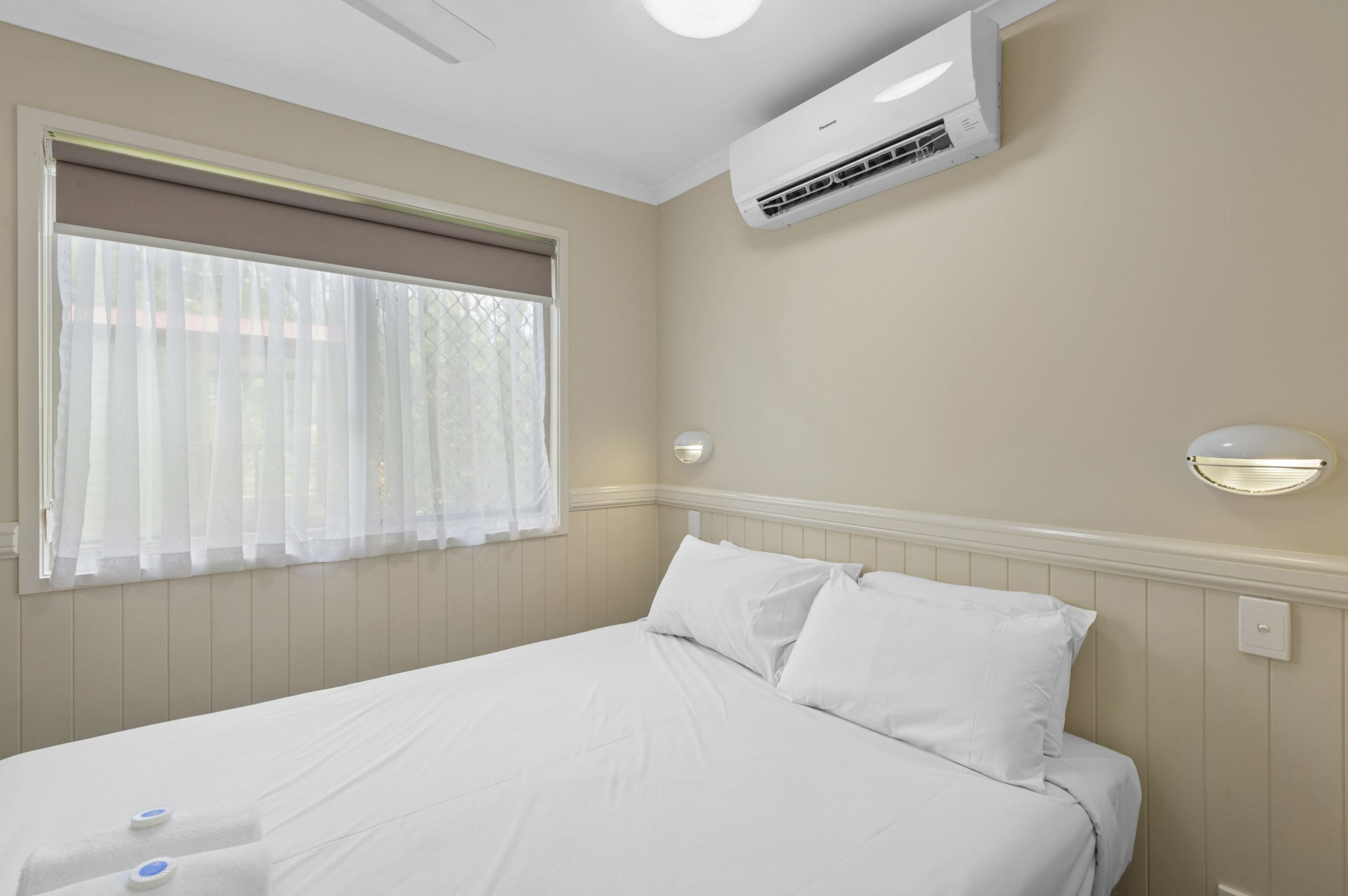 A hotel room with a bed, a window with sheer curtains, an air conditioning unit, and a wall-mounted light fixture.