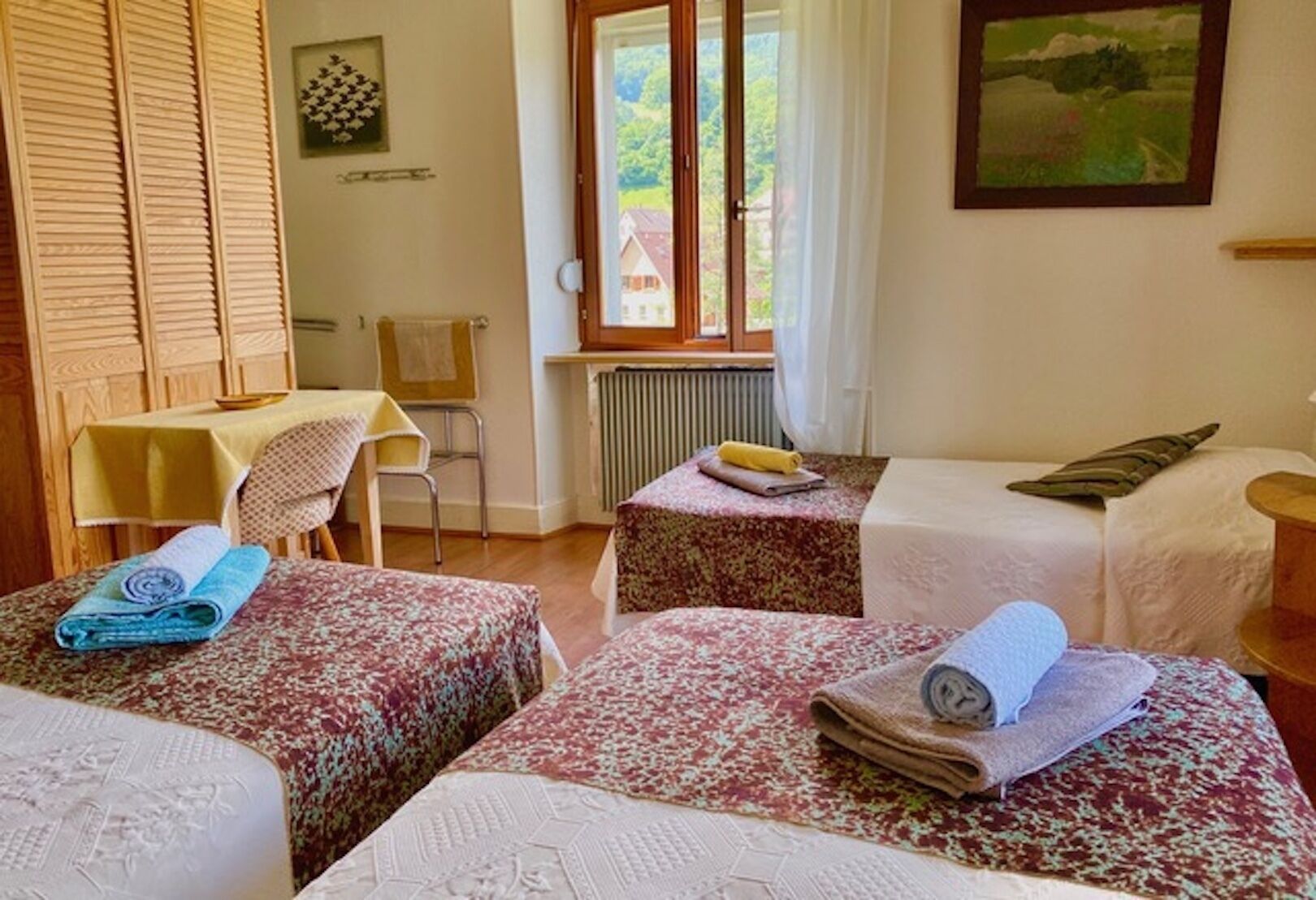 Une chambre avec deux lits, une table, une chaise et une fenêtre donnant sur des maisons.