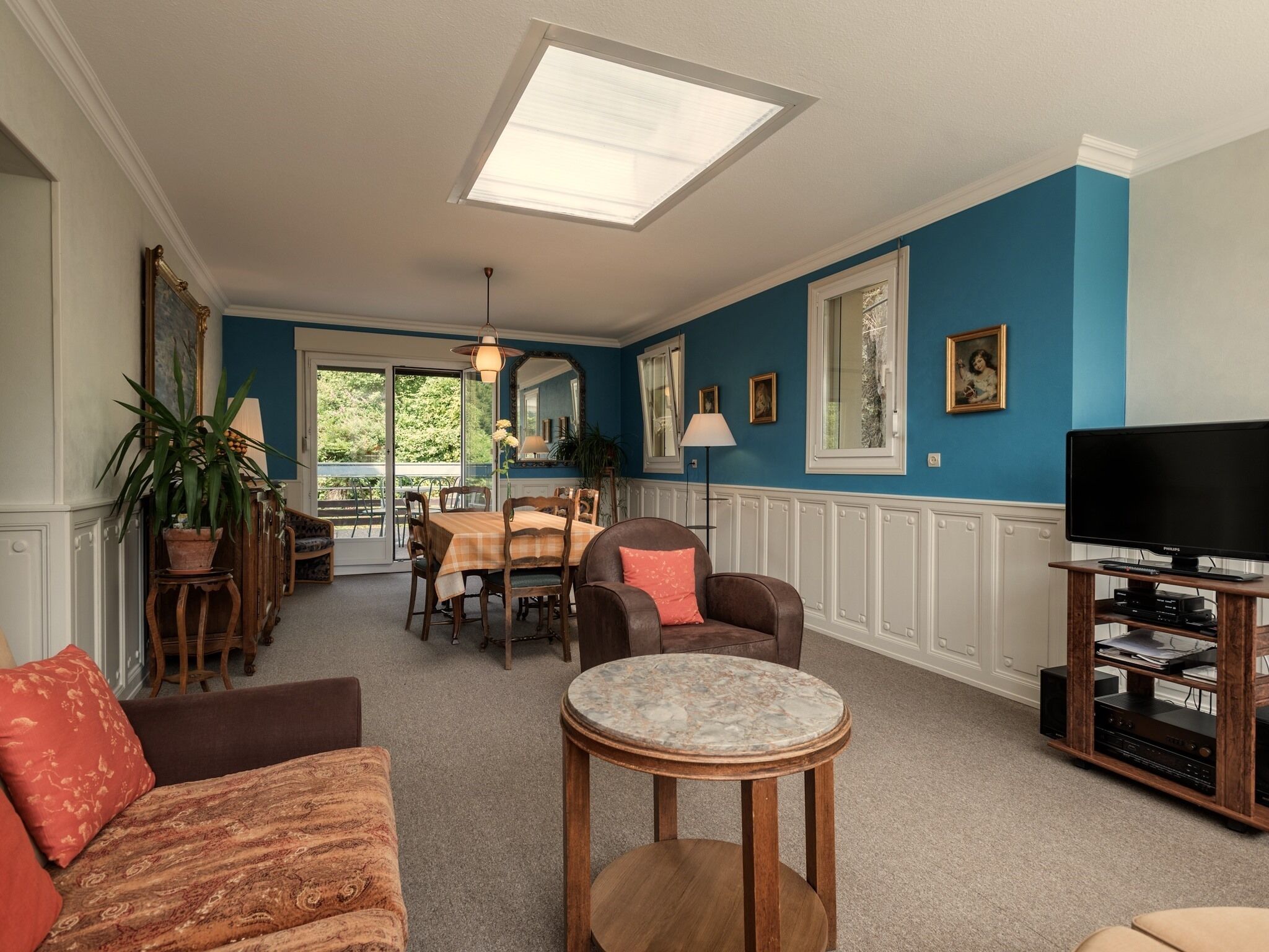 A living room with a blue wall, a dining area with a table and chairs, a sofa, a coffee table, and a television on a wooden stand.