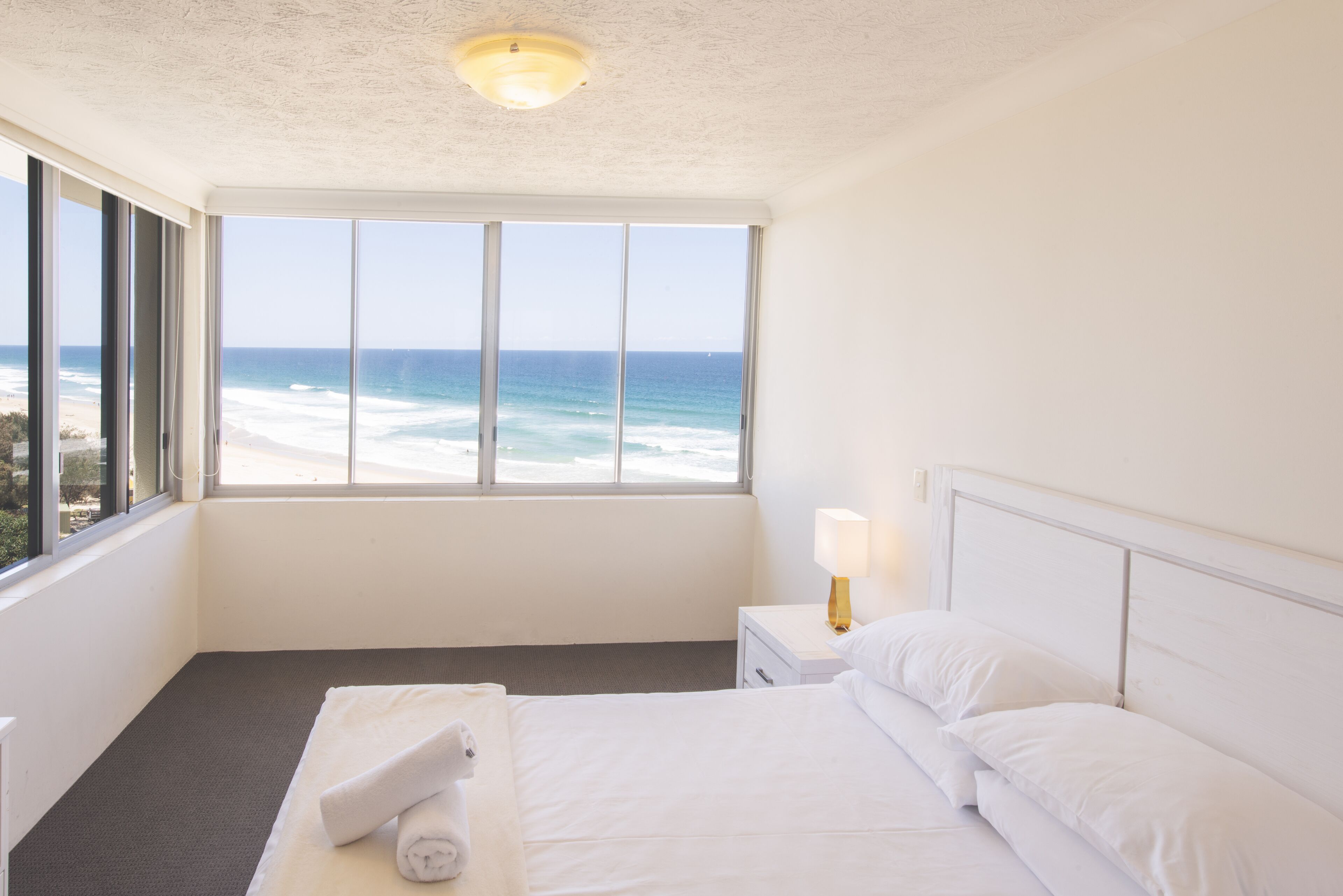 A hotel room with a large window overlooking the ocean, a neatly made bed, a bedside table with a lamp, and a small nightstand.
