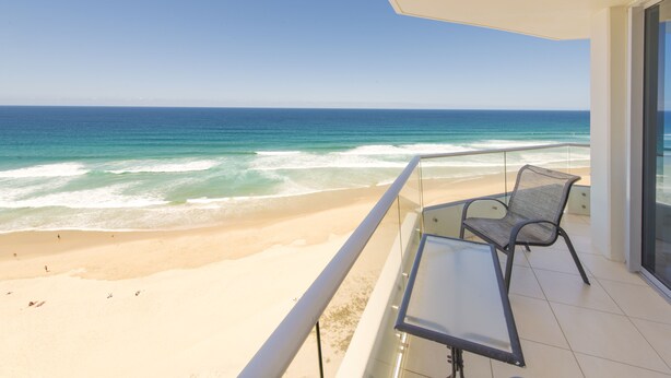 Ein Balkon mit Glasgeländer, der auf einen Strand und das Meer blickt. Auf dem Balkon stehen ein Tisch und zwei Stühle.